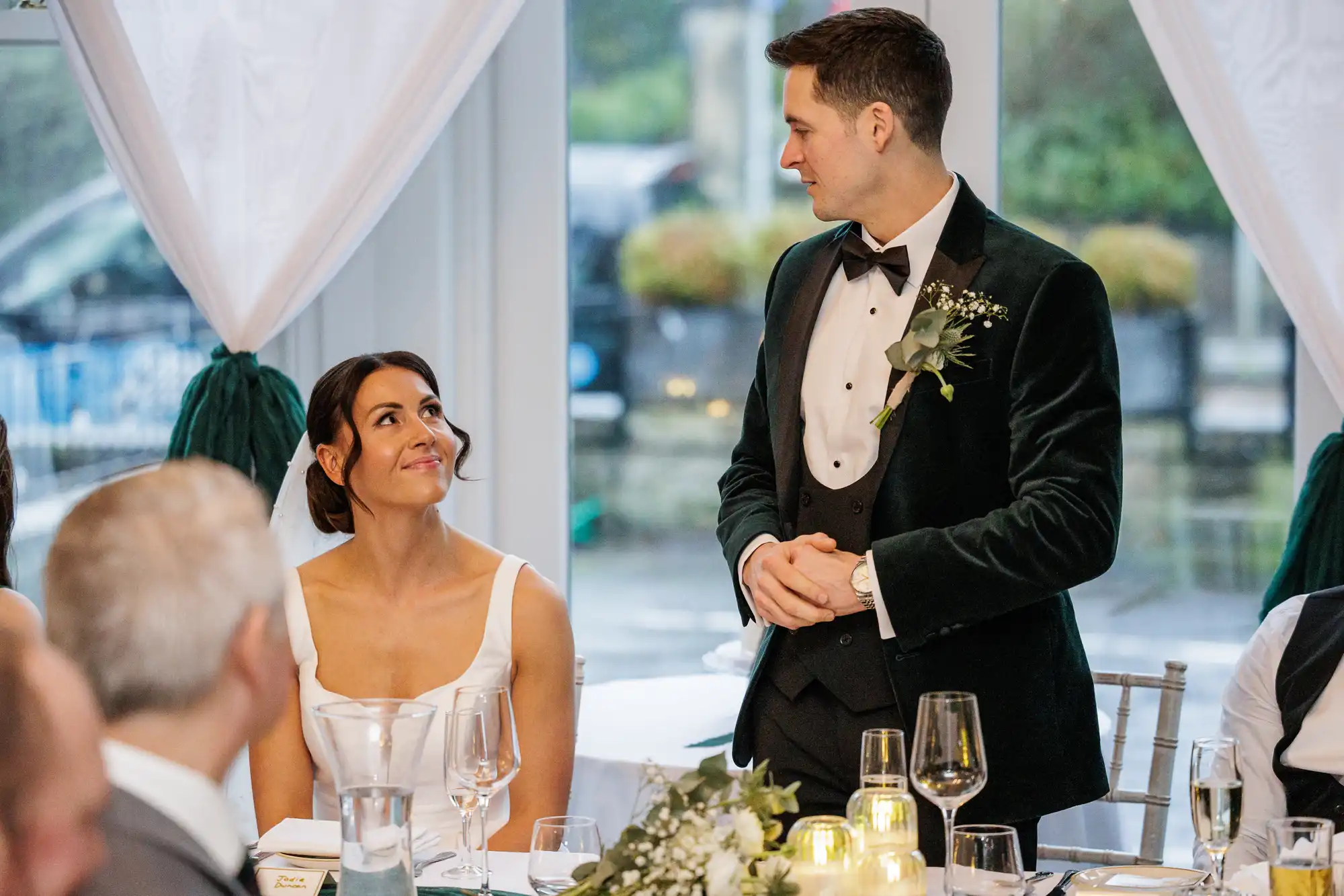 Bride gazes lovingly at groom in a dark green velvet tuxedo. They are at a wedding reception table with floral arrangements and wine glasses.