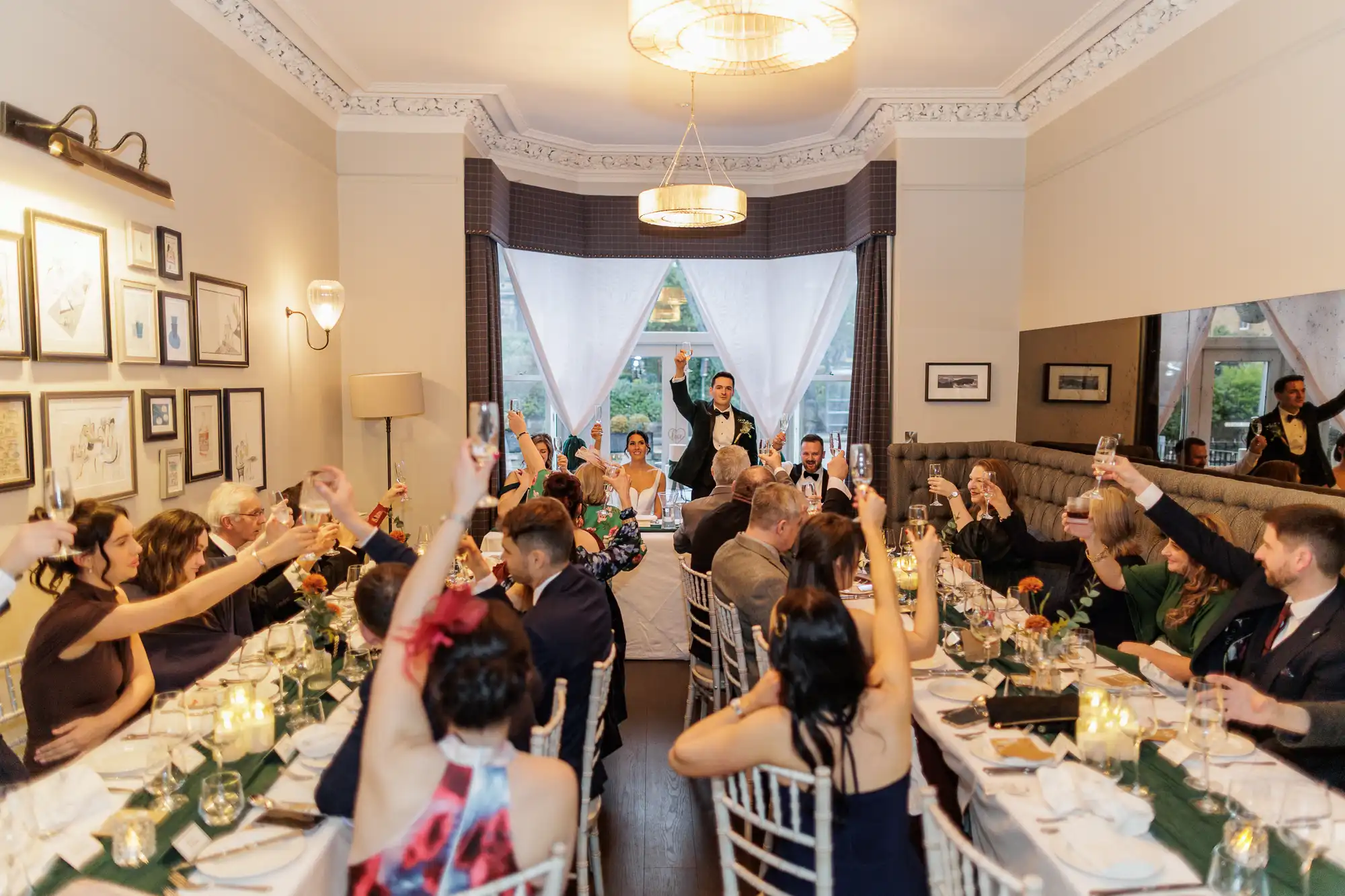 Wedding party and guests raise champagne glasses in a toast during a reception in a warmly lit dining room with framed art on the walls and a large ornate chandelier overhead.