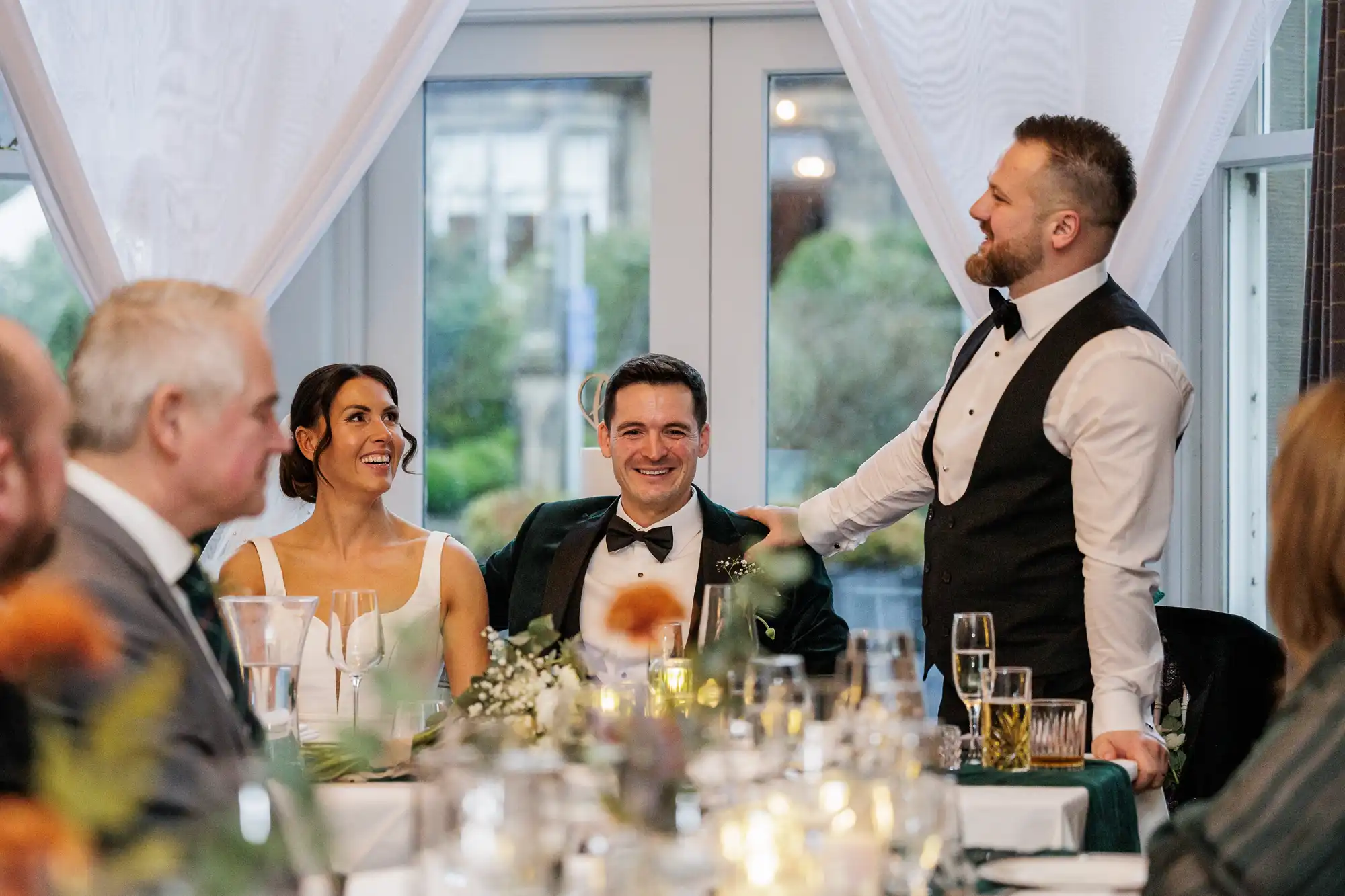 Bride smiles with groom as a man in a waistcoat places a hand on his shoulder at a wedding reception.