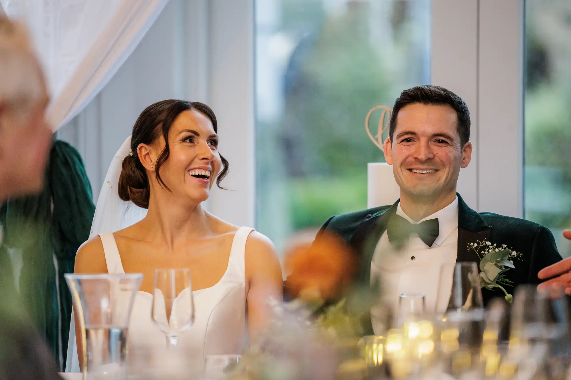 Newlyweds, a bride in a white dress and a groom in a dark green tuxedo, smile broadly while seated at a table during their wedding reception.
