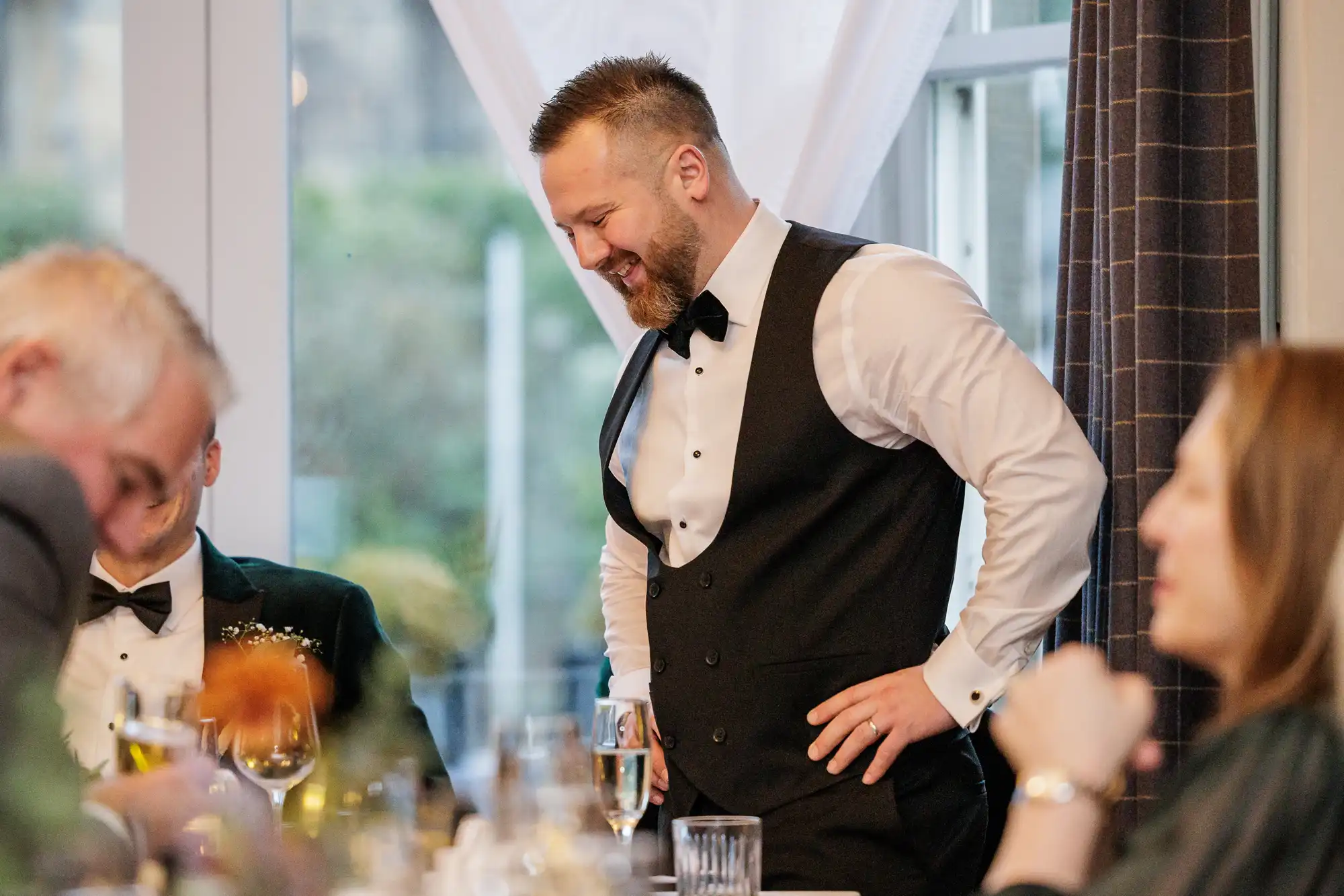 A man with a beard and short hair, wearing a white shirt, black vest, and bow tie, laughs with his hands on his hips.