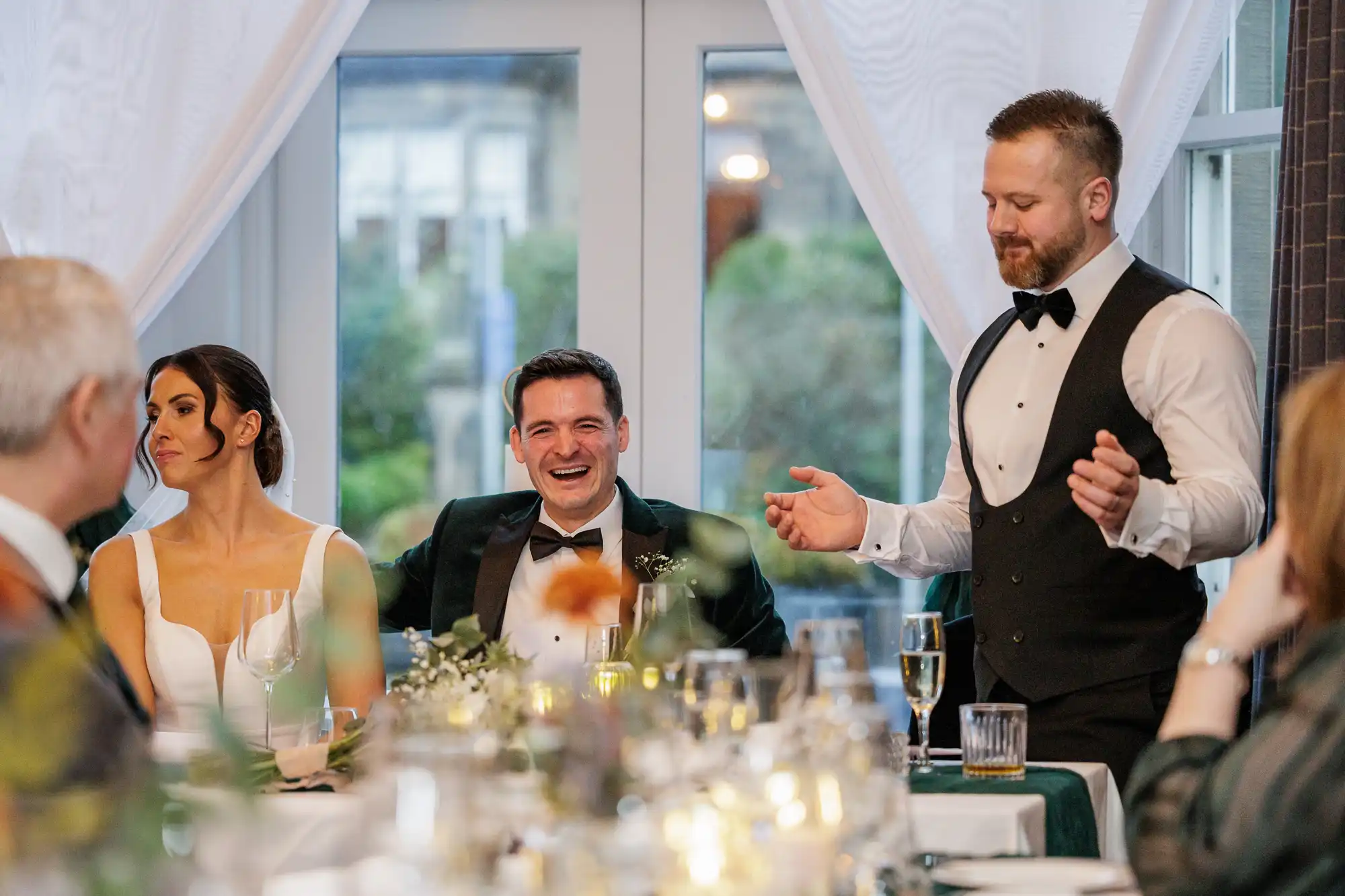 A wedding party sits at a decorated table with a man in a tuxedo laughing heartily and a man in a vest giving a toast.