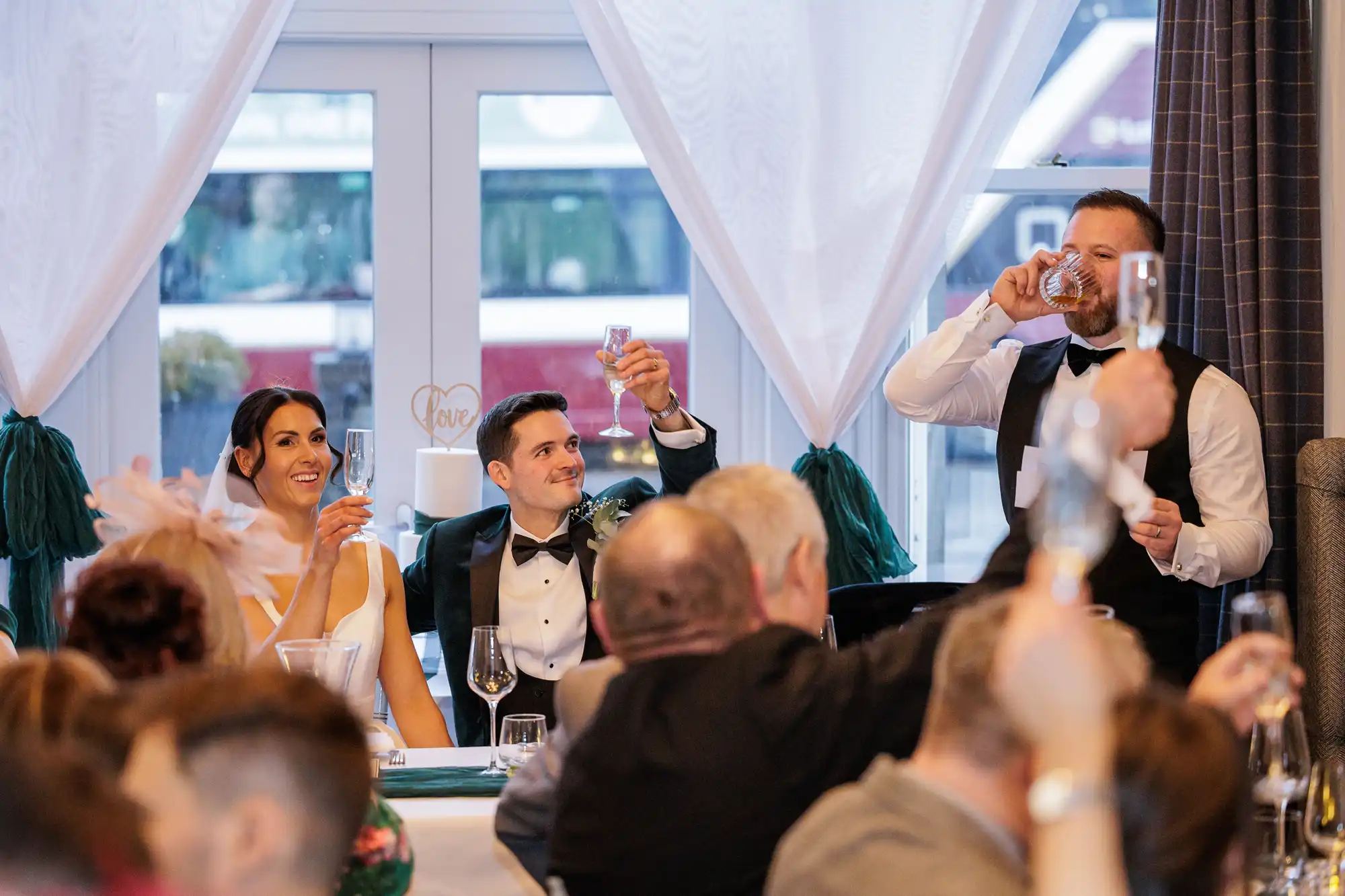 A smiling bride in a white dress holds a champagne flute at a wedding reception. The groom, in a velvet jacket, raises his glass.