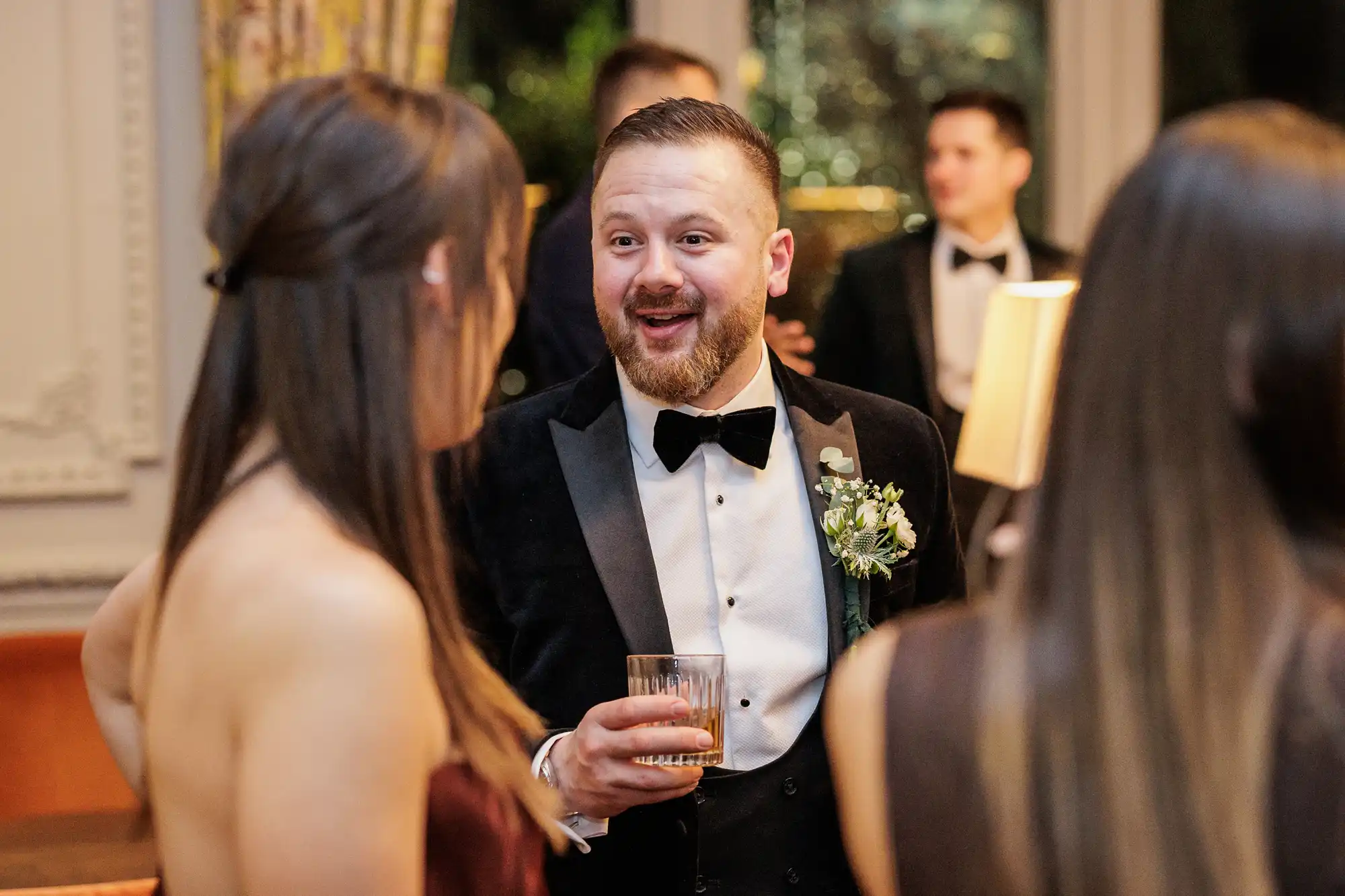 A man in a tuxedo smiles, holding a drink and talking to a woman in a maroon dress. He has a boutonnière on his lapel.
