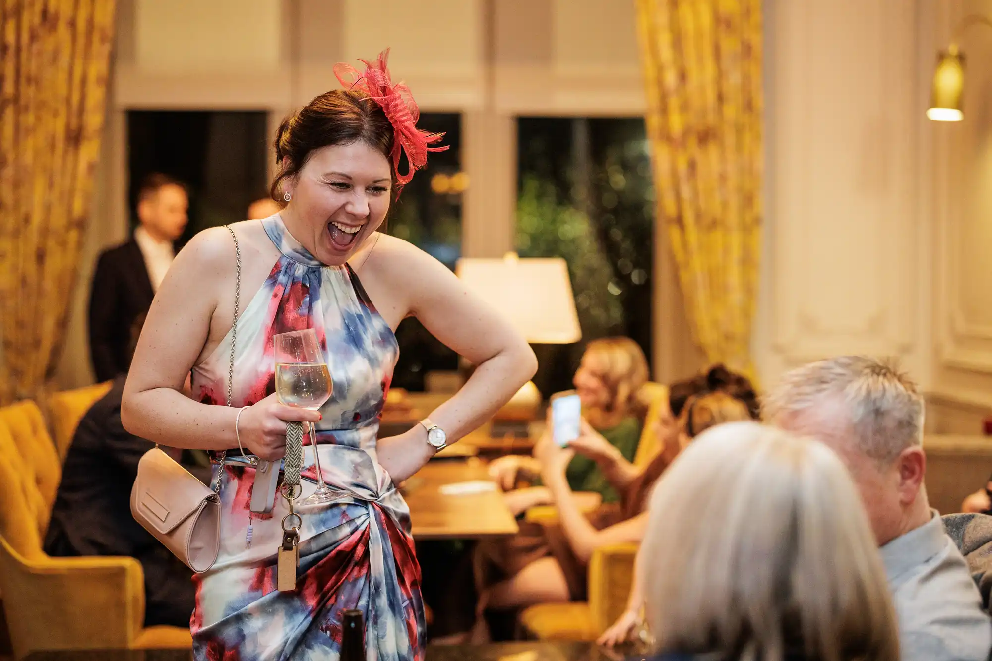 A woman in a colorful dress and red fascinator laughs with her hand on her hip, holding a glass of white wine. A blush pink purse hangs from her shoulder.