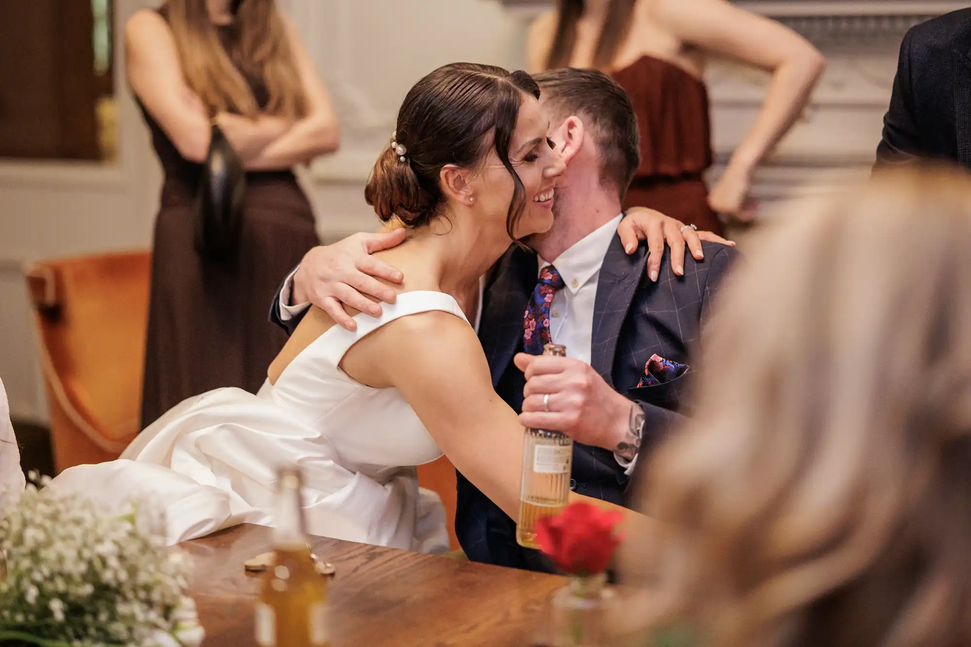 A bride in a white dress with her arms around a groom in a navy suit. They are both smiling and hugging. The groom holds a bottle.