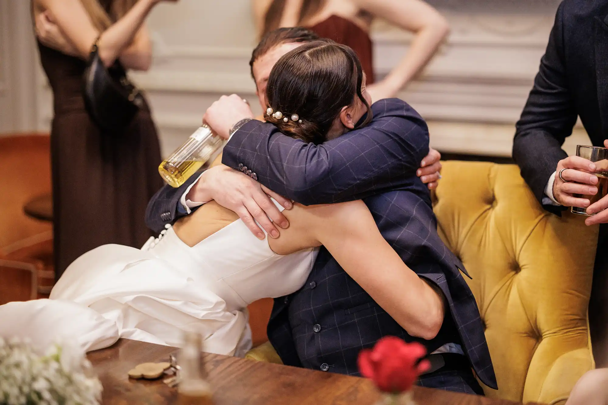 A couple hugs tightly, the man holding a bottle of liquor and the woman in a white wedding dress, adorned with pearl hairpins. A man in a suit holds a drink in the background.