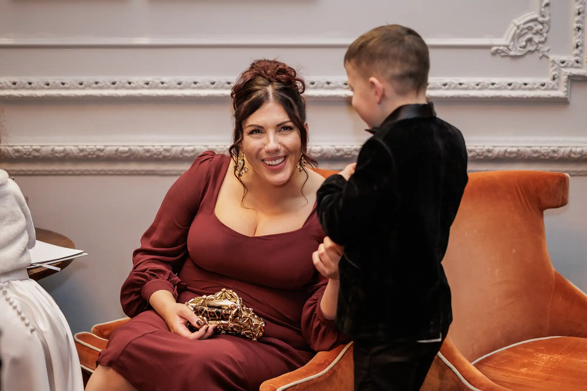A woman in a maroon dress laughs as a young boy in a black velvet suit talks to her. They are sitting on an orange velvet chair in a room with ornate white molding.