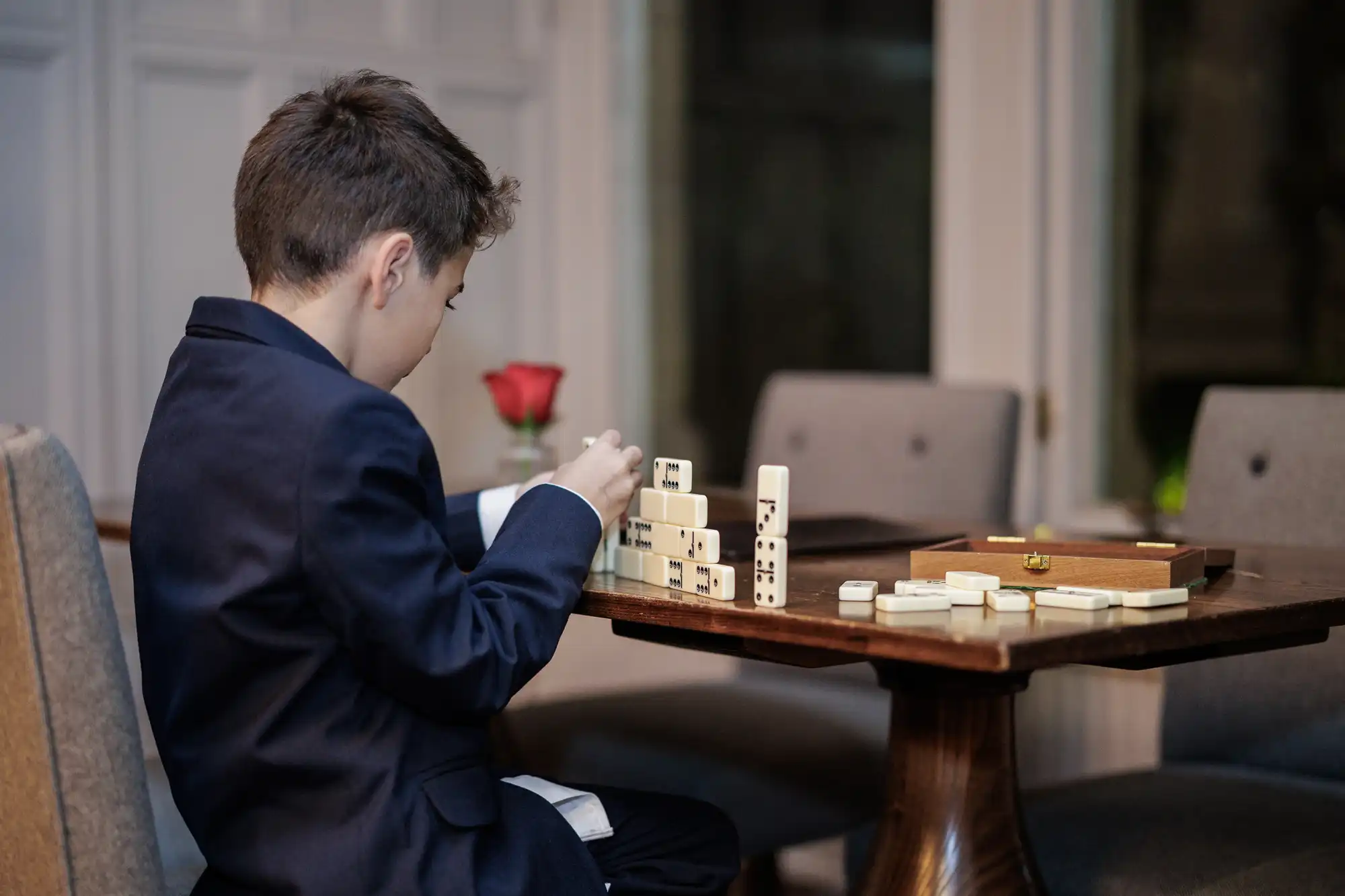 A boy in a dark suit plays dominoes at a wooden table, stacking the tiles in a pyramid and on a single line.