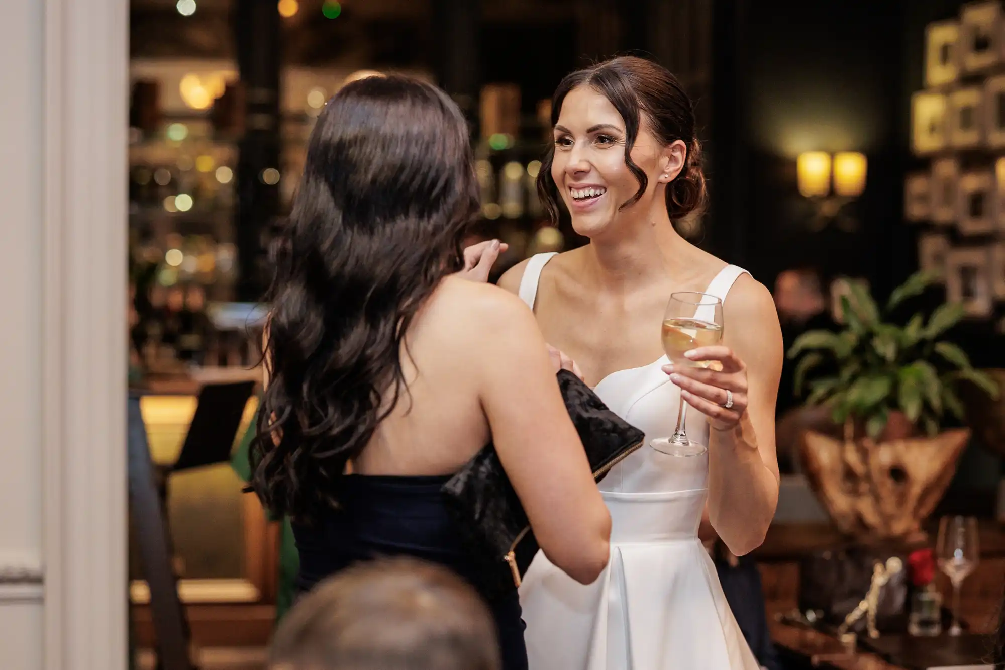 Bride in a white dress laughing with a guest holding a champagne flute. She wears a ring on her left hand.