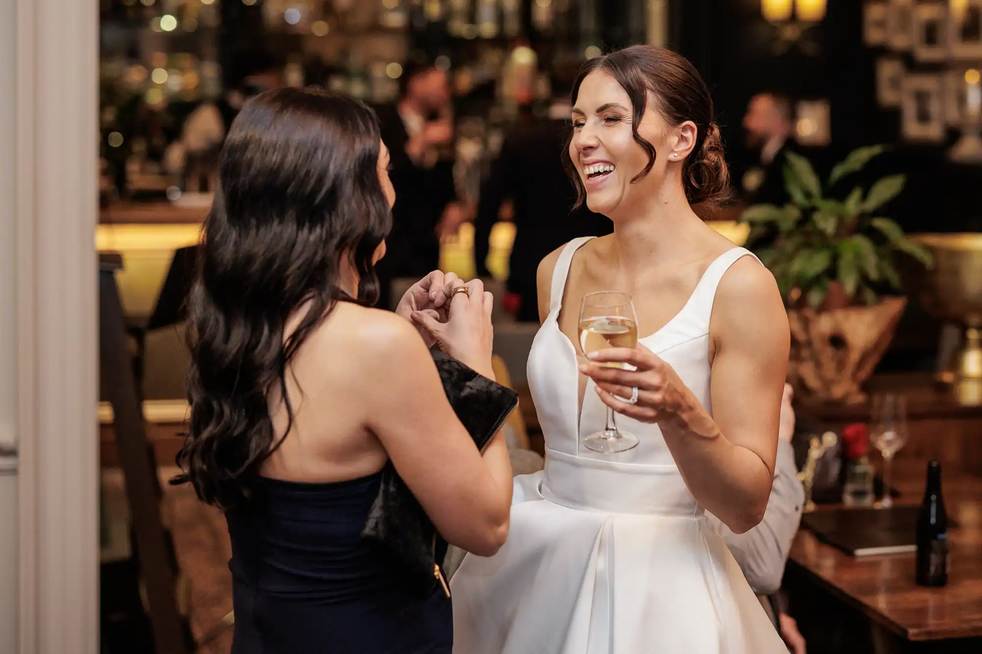 Two women in formal attire converse in a dimly lit venue. The woman on the right, wearing a white dress and holding a wine glass, laughs heartily. The woman on the left, in a dark dress, looks towards her with a clutch bag.