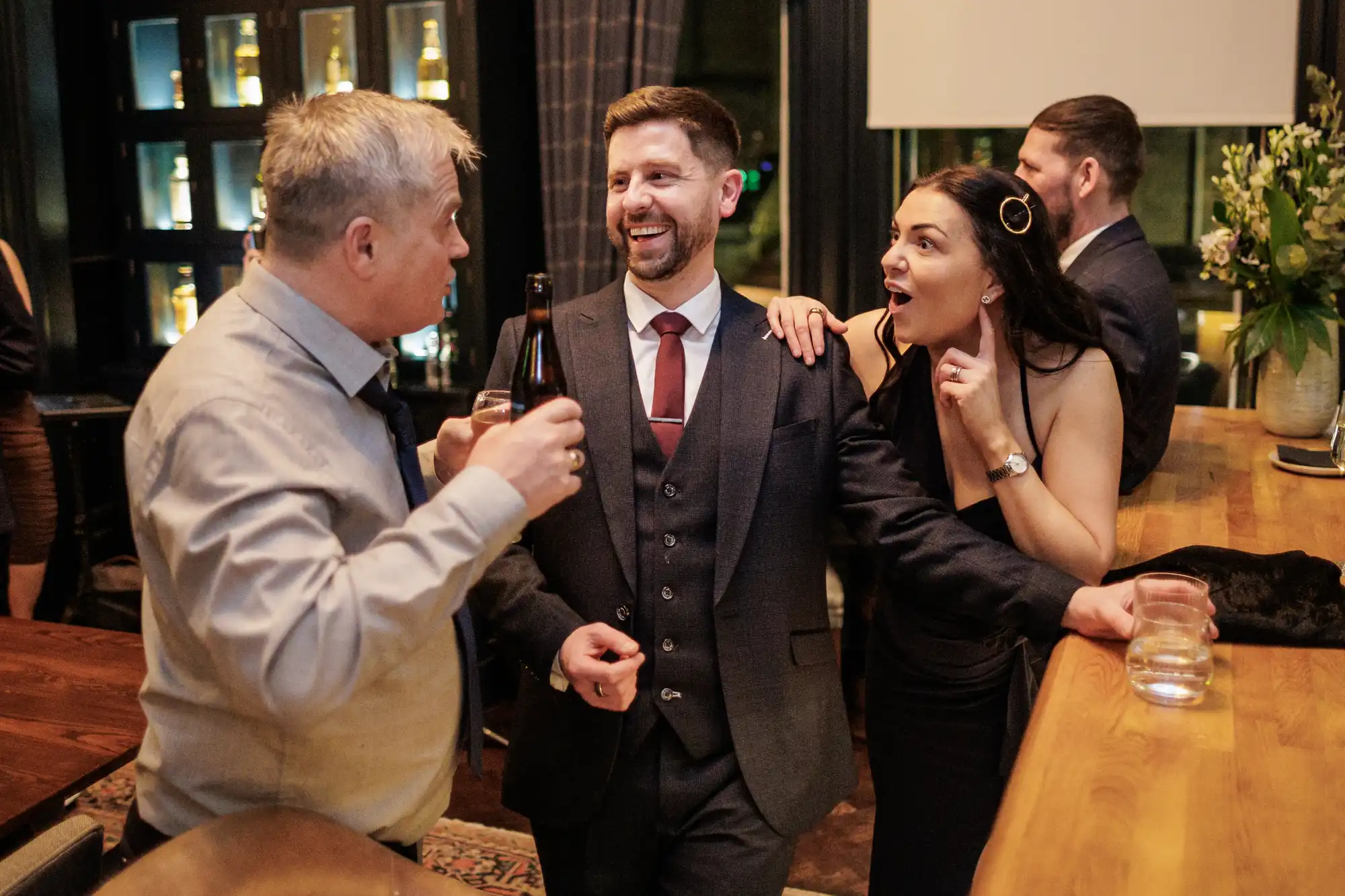 Three people in formal attire laugh and talk at a bar. The man in the center wears a suit, the man on the left a shirt and tie, and the woman on the right a black dress.