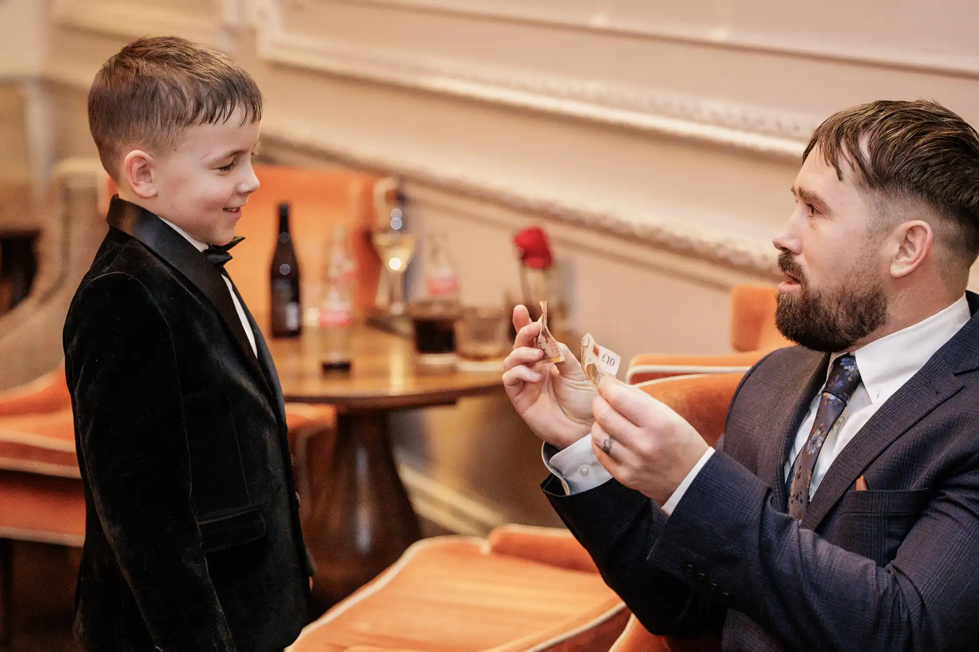 A boy in a tuxedo smiles at a man in a suit who is holding up two ten-pound notes.
