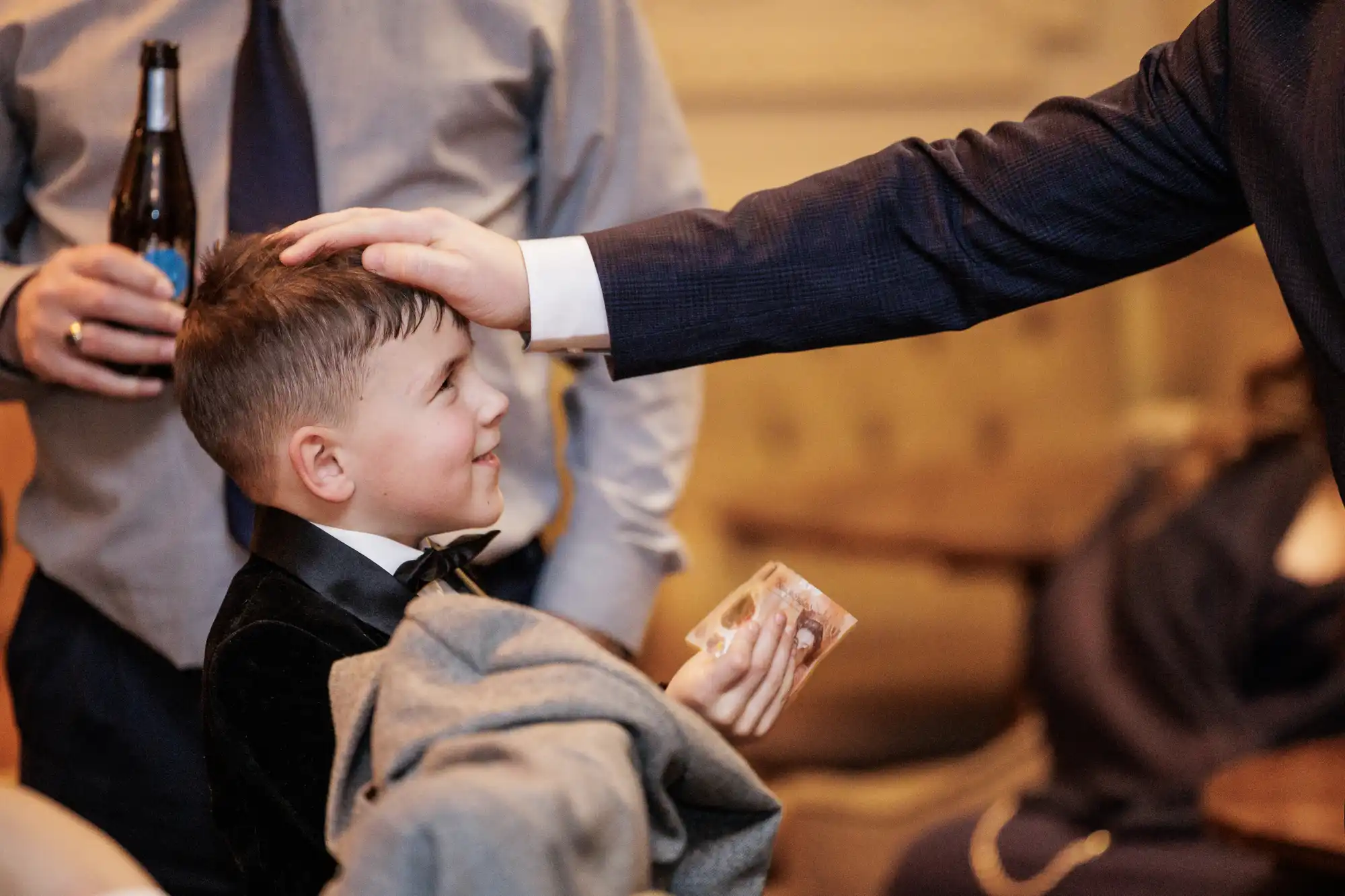 A smiling boy in a tuxedo receives money from a man in a suit. Another man in the background holds a bottle.