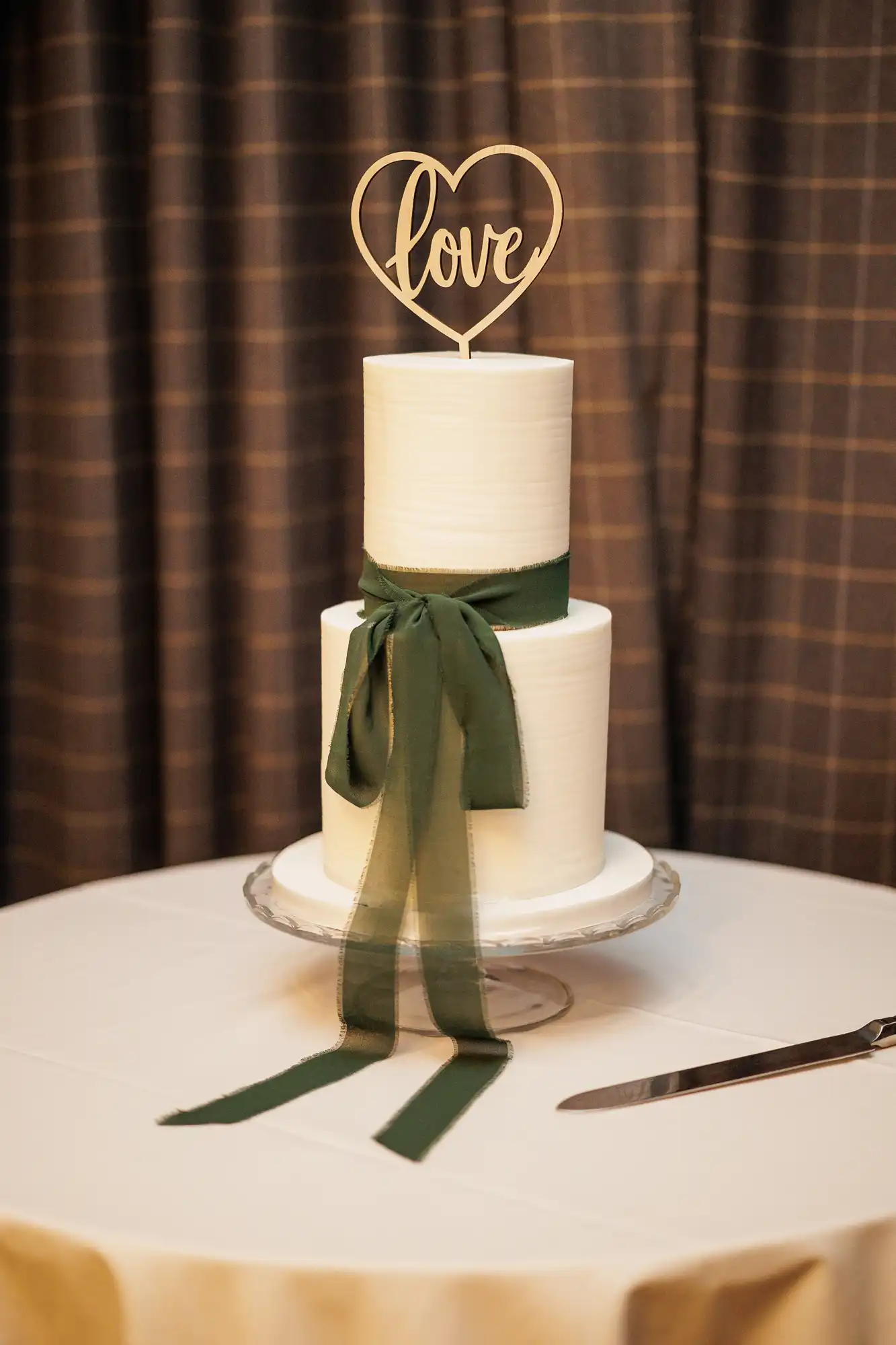 A two-tiered white cake with a wooden "love" heart topper is decorated with a dark green ribbon. A cake knife rests on the table next to it.