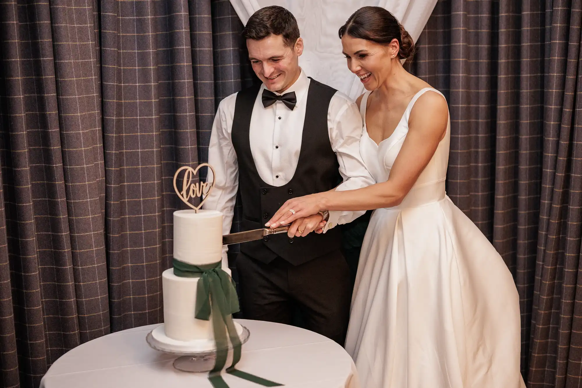 A bride and groom smile as they cut a two-tiered white wedding cake. The cake is decorated with a wooden "love" heart topper and a green ribbon. The couple stands in front of dark, checkered curtains.