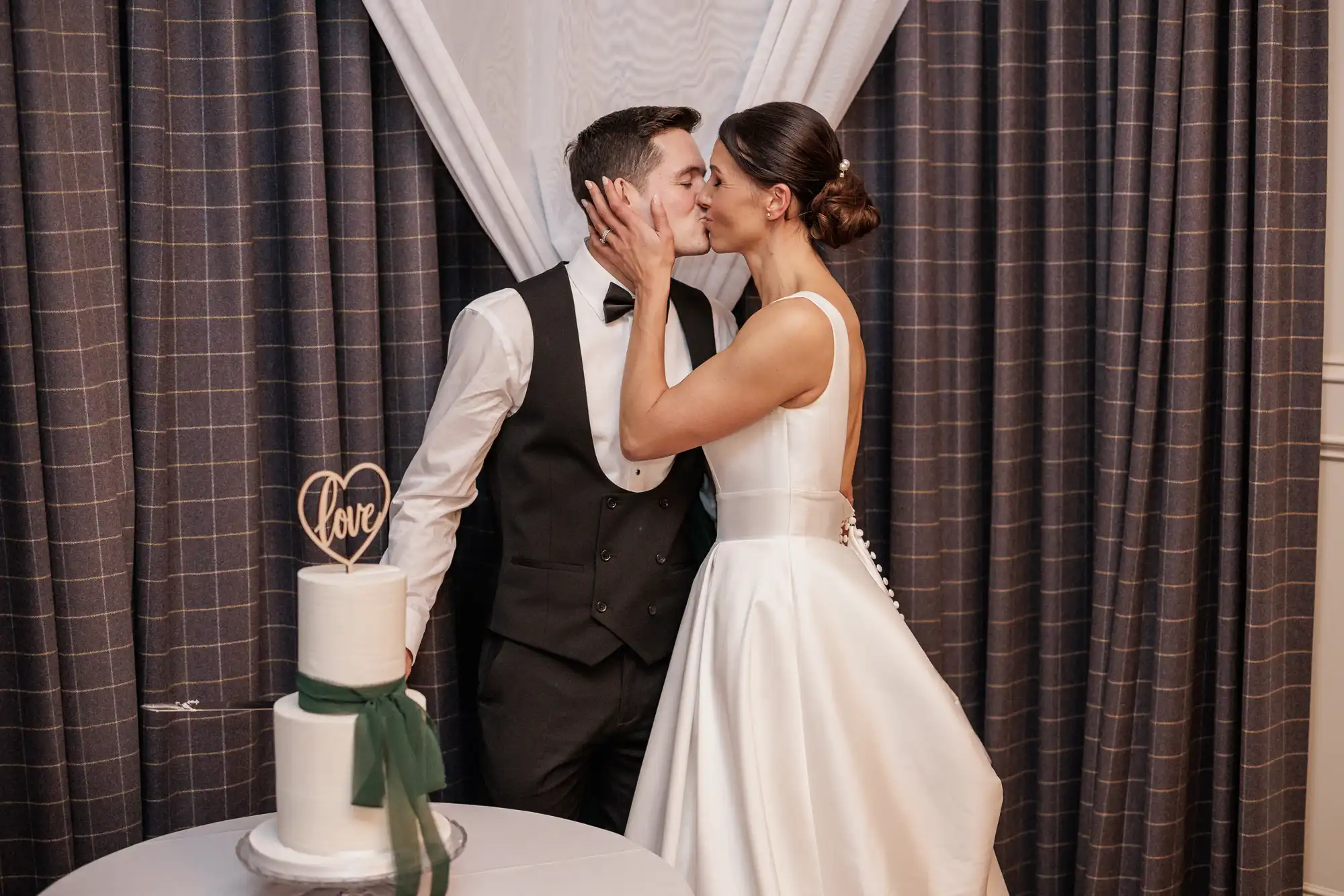 A bride in a white gown and a groom in a tuxedo share a kiss next to a two-tiered cake adorned with a "love" cake topper and a green ribbon.