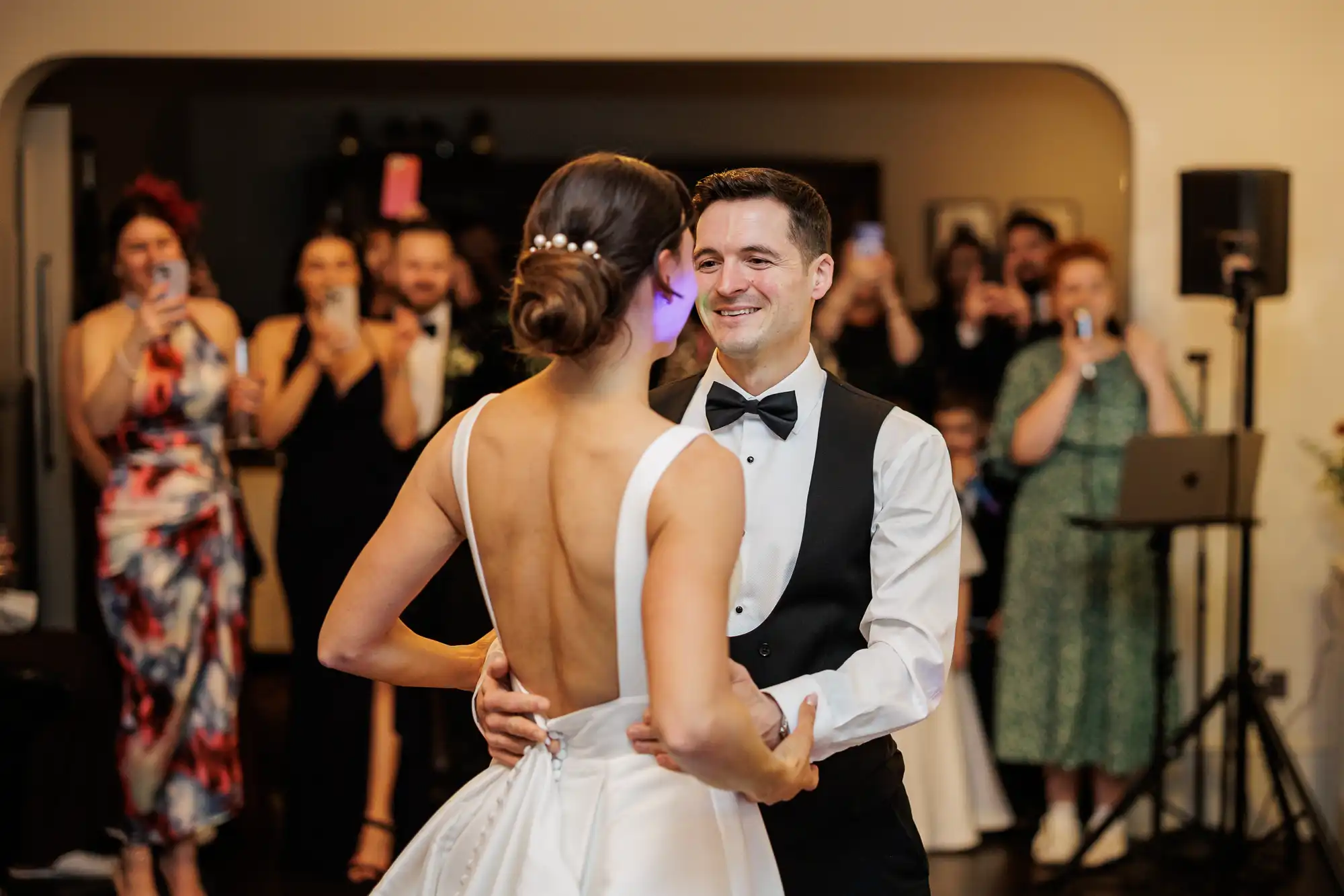 A groom in a vest and bow tie dances with his bride in a low-back dress. Guests in the background hold up phones to capture the moment.