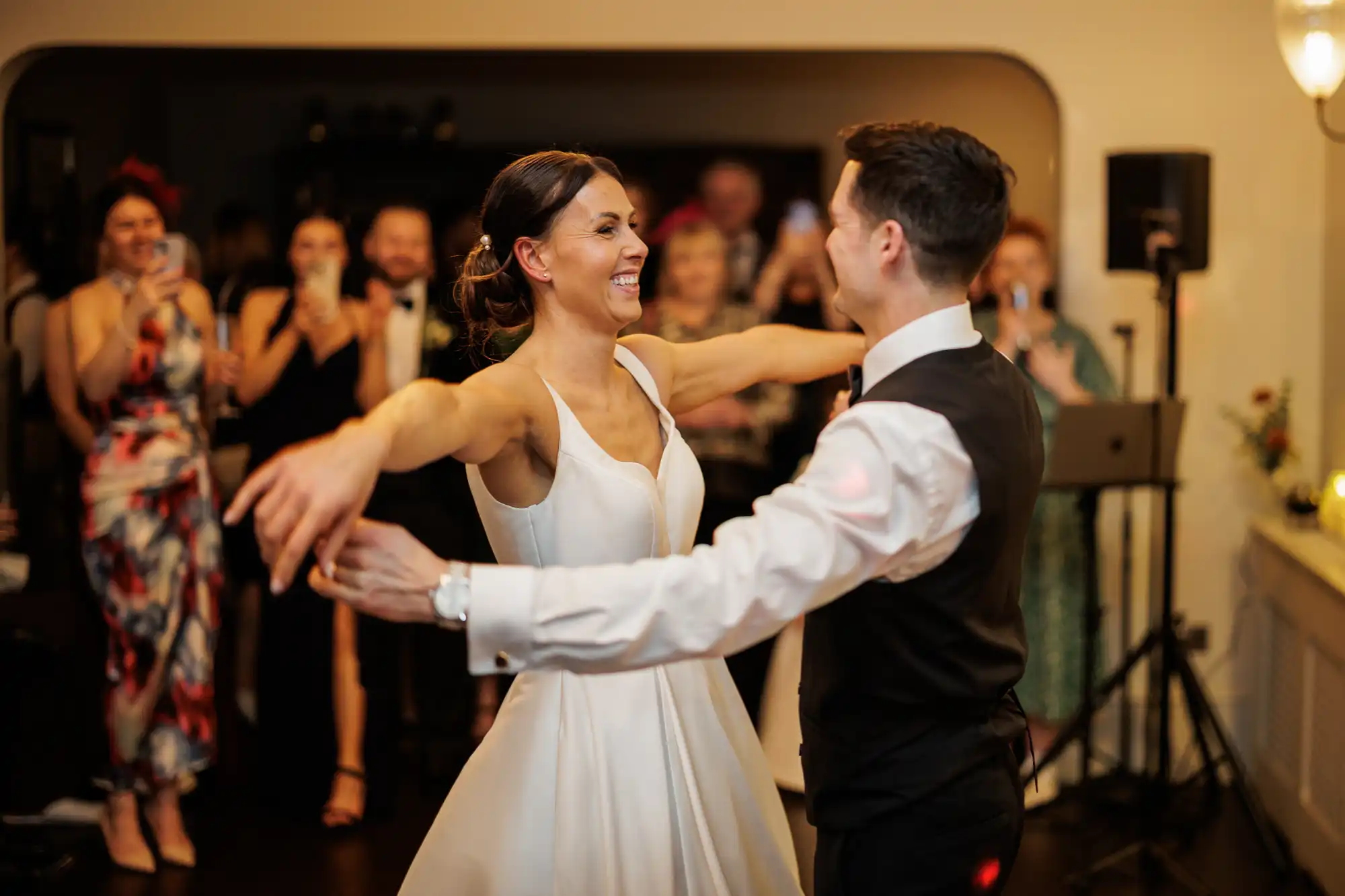 A smiling bride in a white dress dances with her groom at their wedding reception, surrounded by guests.
