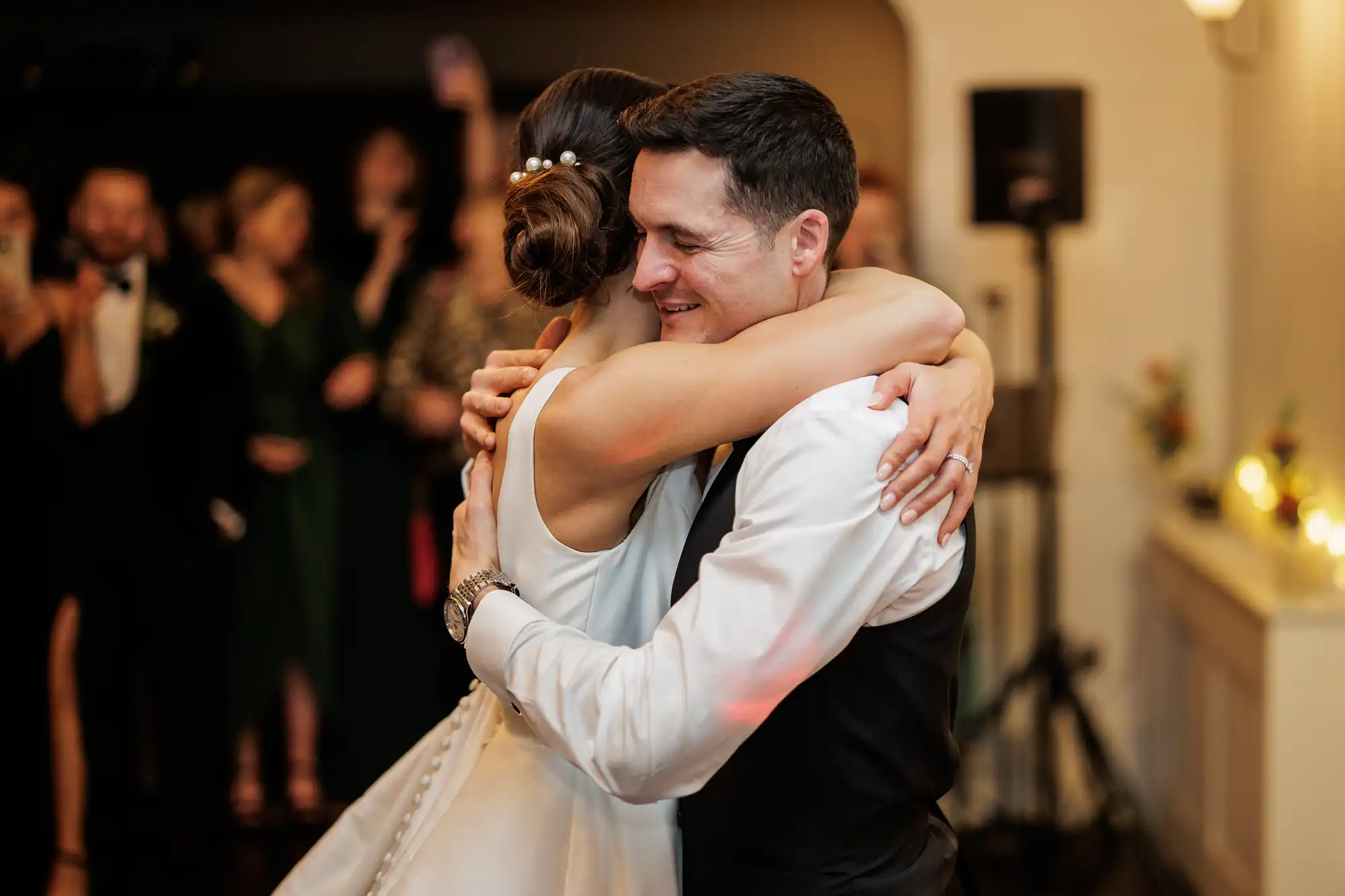 A groom embraces his bride, both smiling, at their wedding reception. Guests are visible blurred in the background.