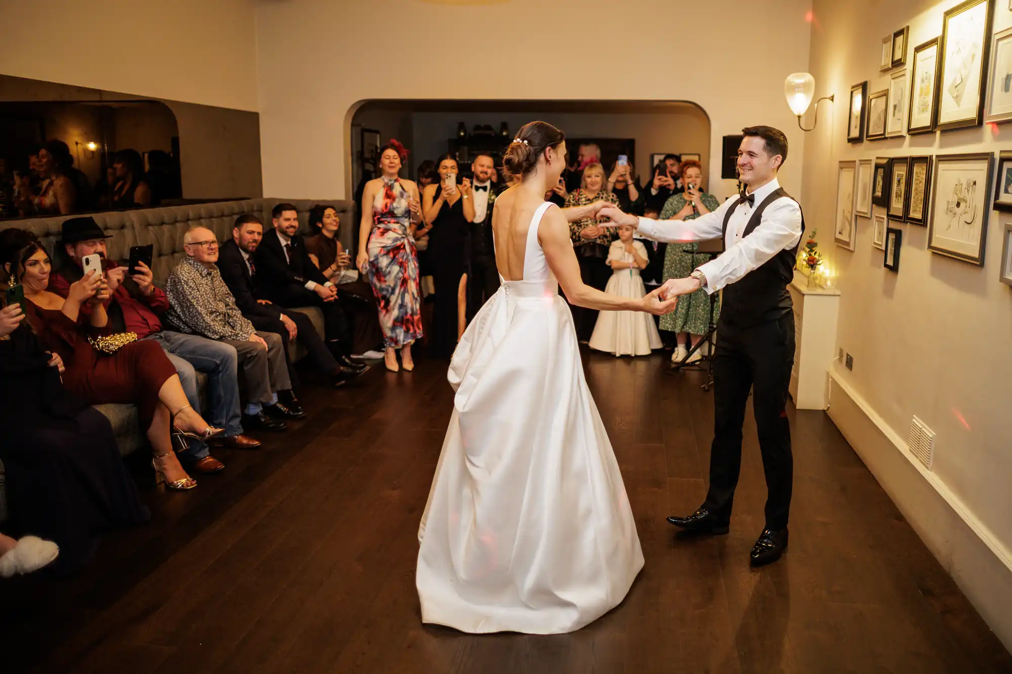A bride in a white gown and groom in a tuxedo dance as guests watch and take photos at a wedding reception.