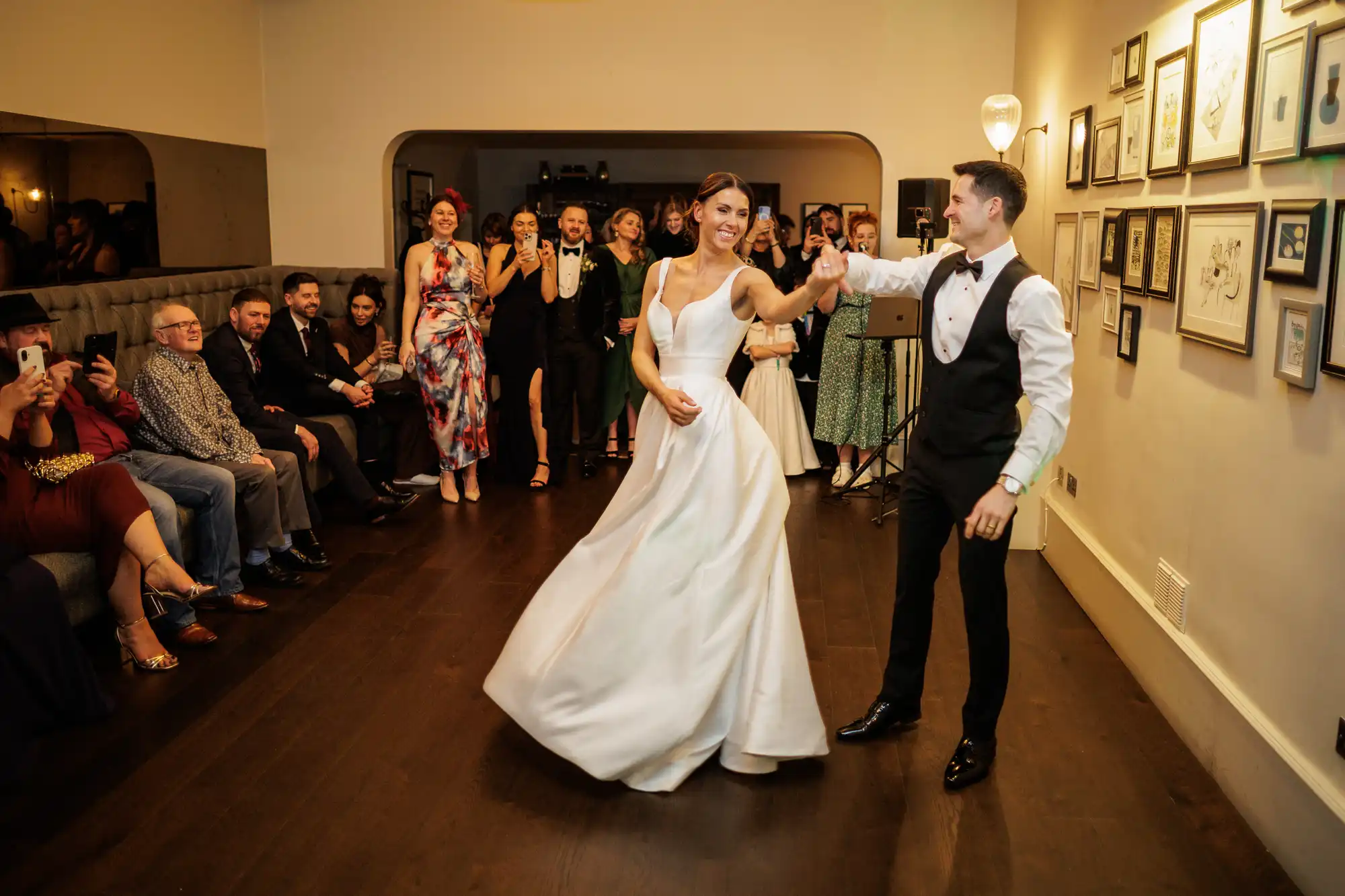 A bride and groom dance in a room with guests watching. The bride wears a white dress, and the groom wears a tuxedo.