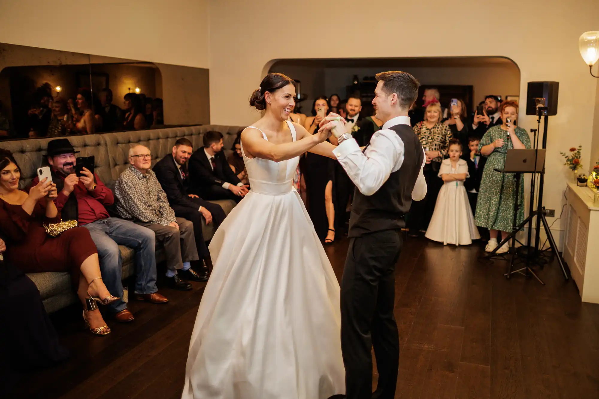 Newlyweds dance together at a wedding reception. The bride wears a white satin gown and the groom a vest and tie. Guests are seated and standing around them, some holding up phones to capture the moment.