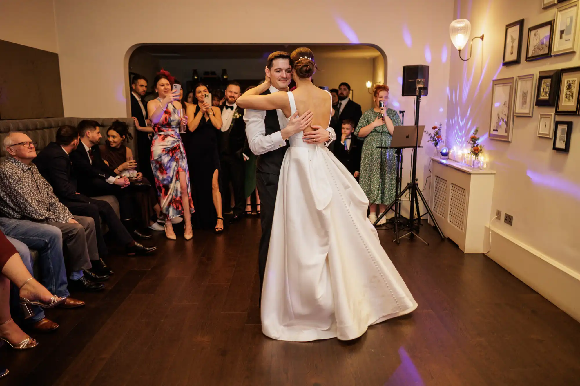 A smiling groom in a dark suit shares his first dance with a bride in a backless white gown, while wedding guests watch, clap, and take photos in a warmly lit reception venue with purple mood lighting.