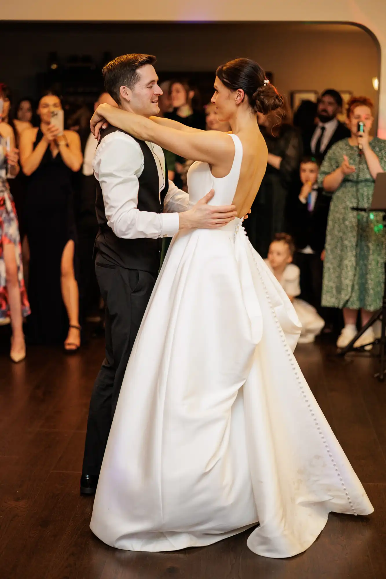 A groom in a suit and a bride in a white ballgown dance together at their wedding reception, surrounded by guests.