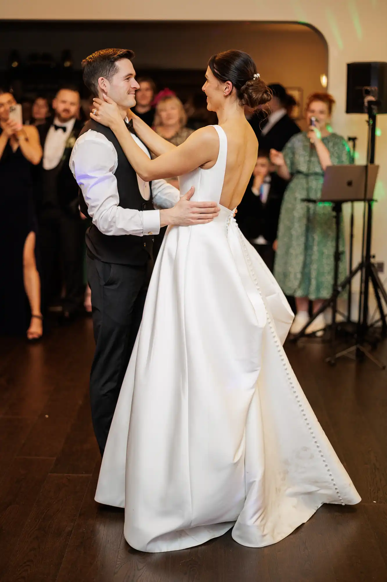A bride in a white satin gown dances with her groom. She looks over her shoulder, her hair in a bun. He smiles at her, holding her close.