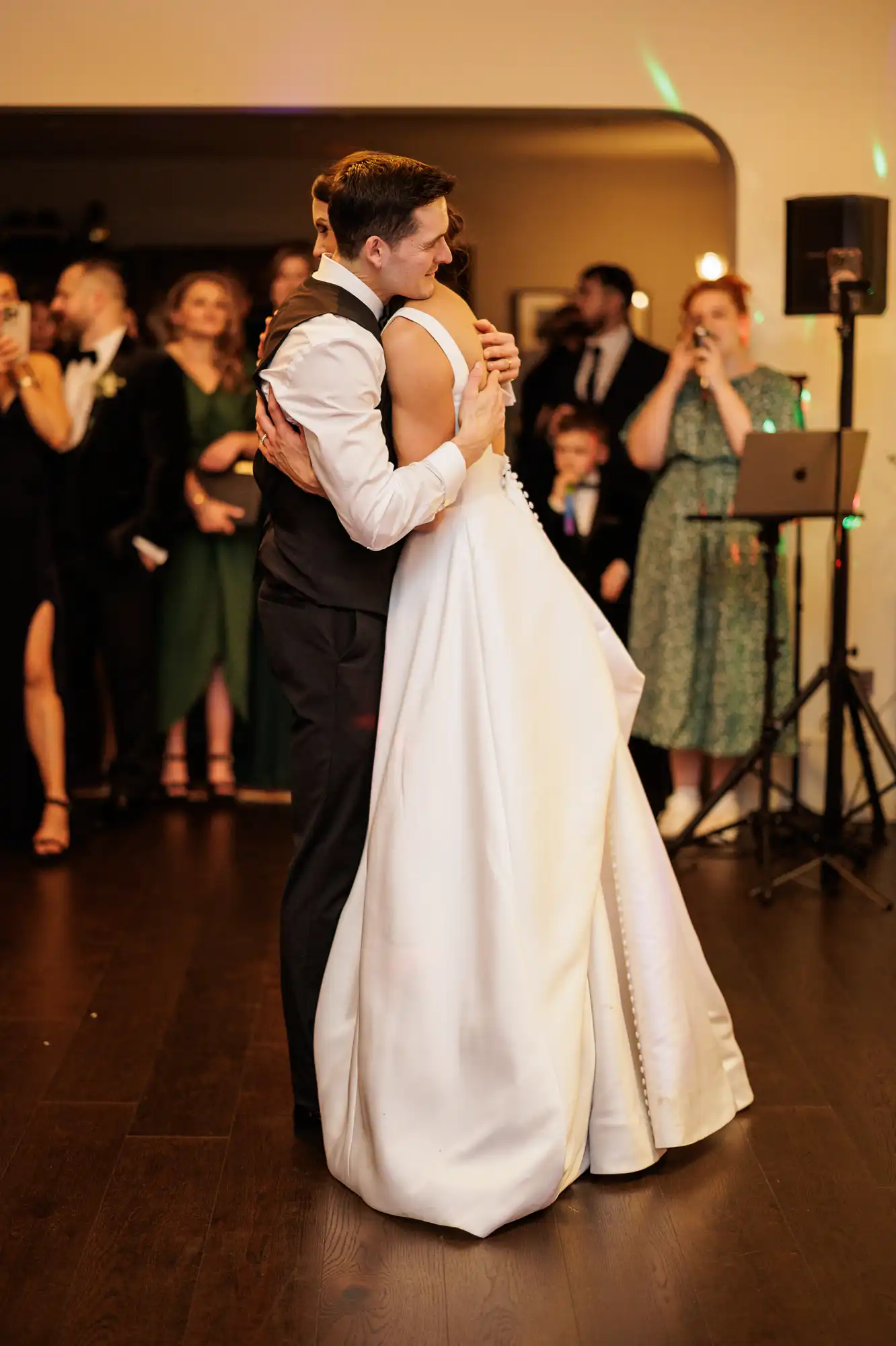 A groom in a suit and vest hugs his bride, who wears a white wedding dress, while guests watch at a reception.