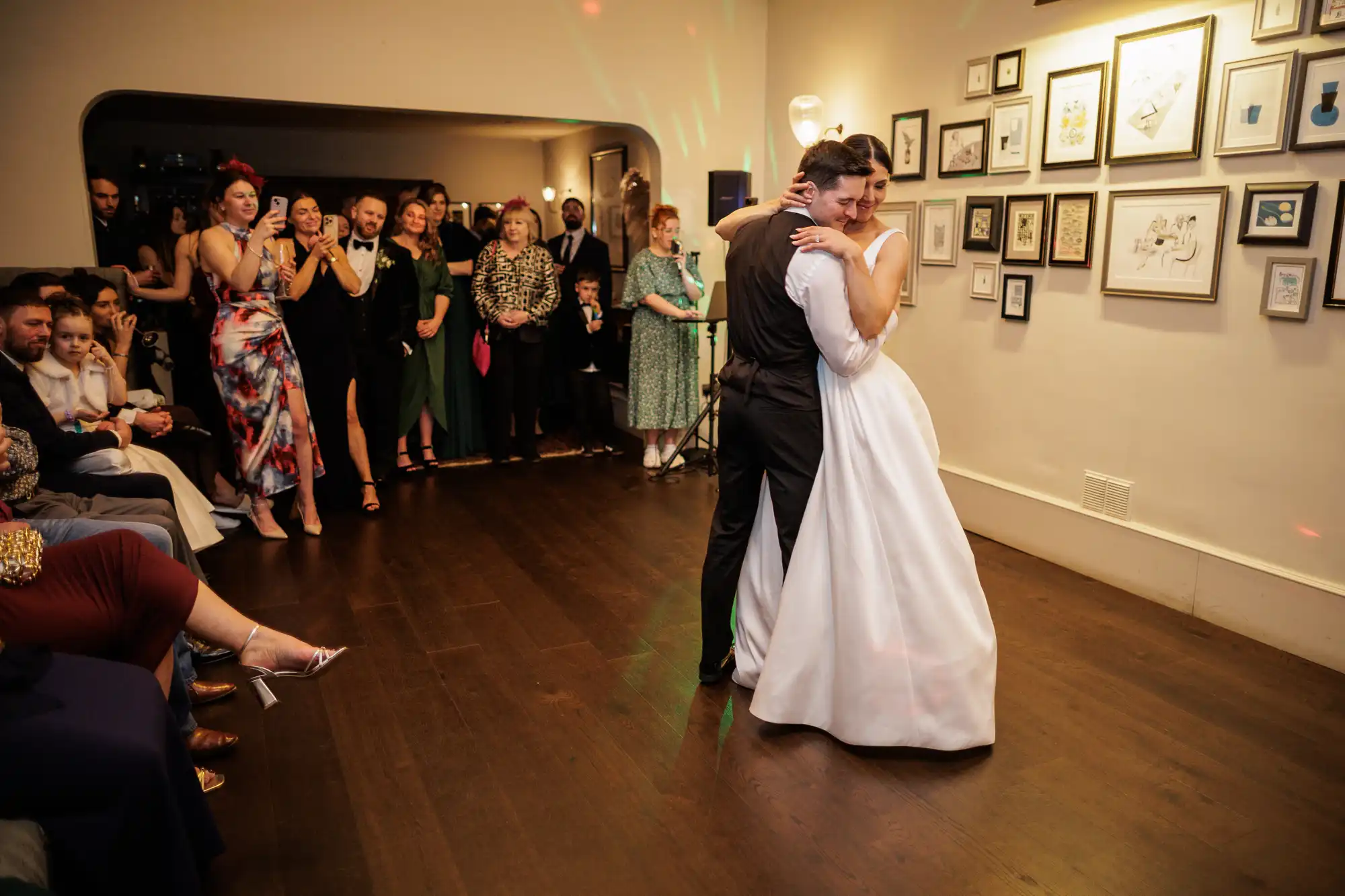 A bride in a white dress and groom in a black suit dance closely at their wedding reception, with guests watching in the background.