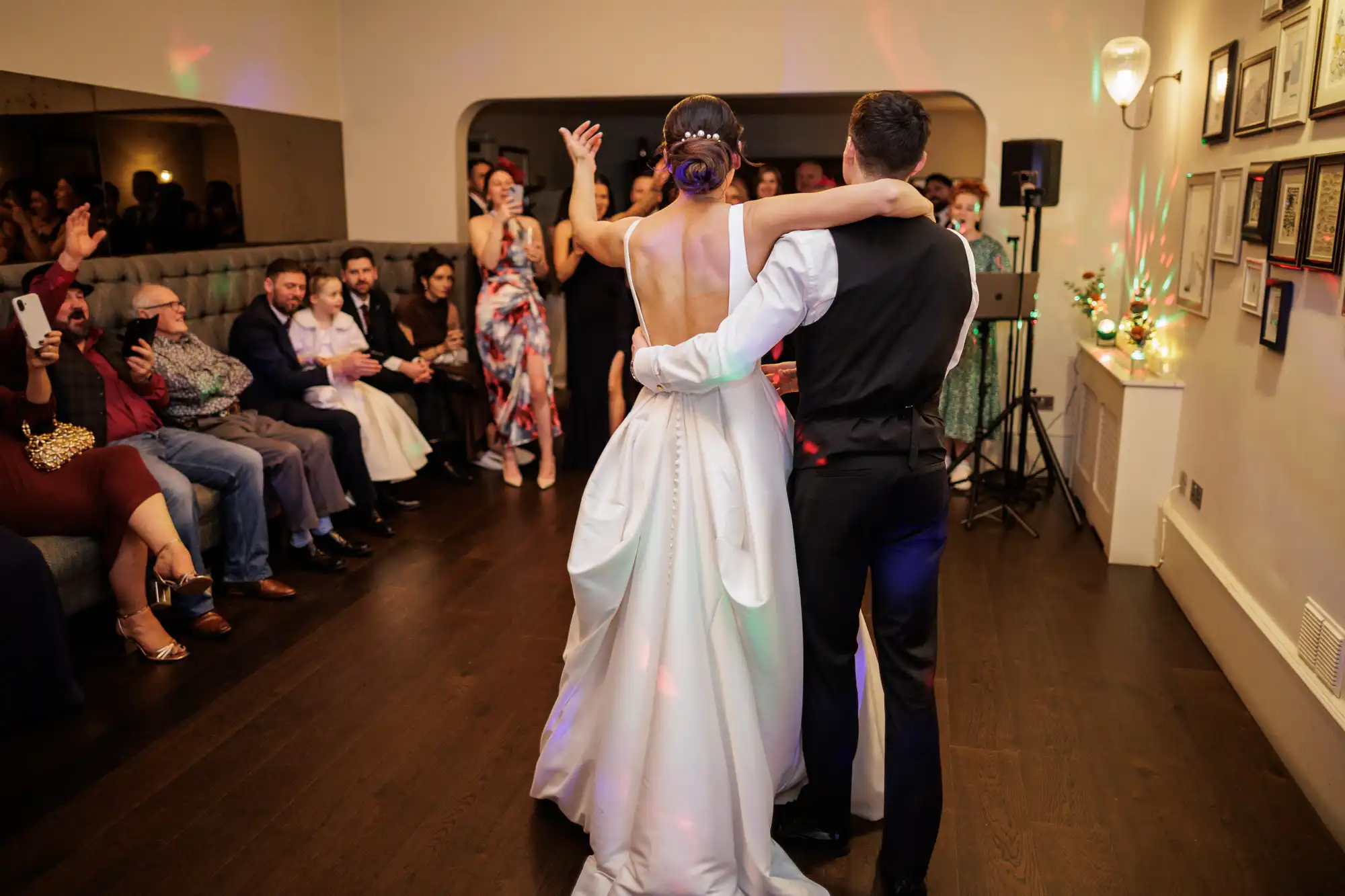 Bride in a backless white gown and groom in a dark suit share their first dance, seen from behind. Cheering wedding guests watch and film from seating areas. Colorful disco lights illuminate the indoor venue.