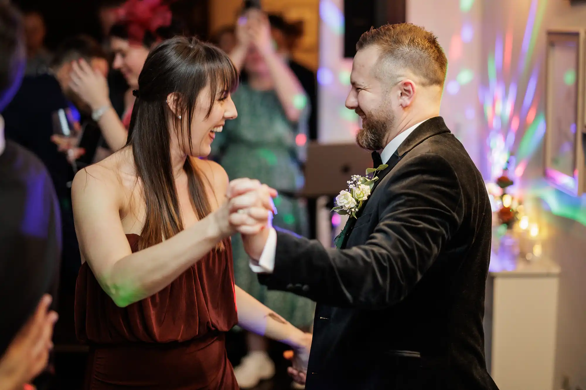 A happy couple dances at a wedding reception, bathed in colorful lights. The bride in a rust-colored dress laughs, while the groom in a velvet suit smiles back.