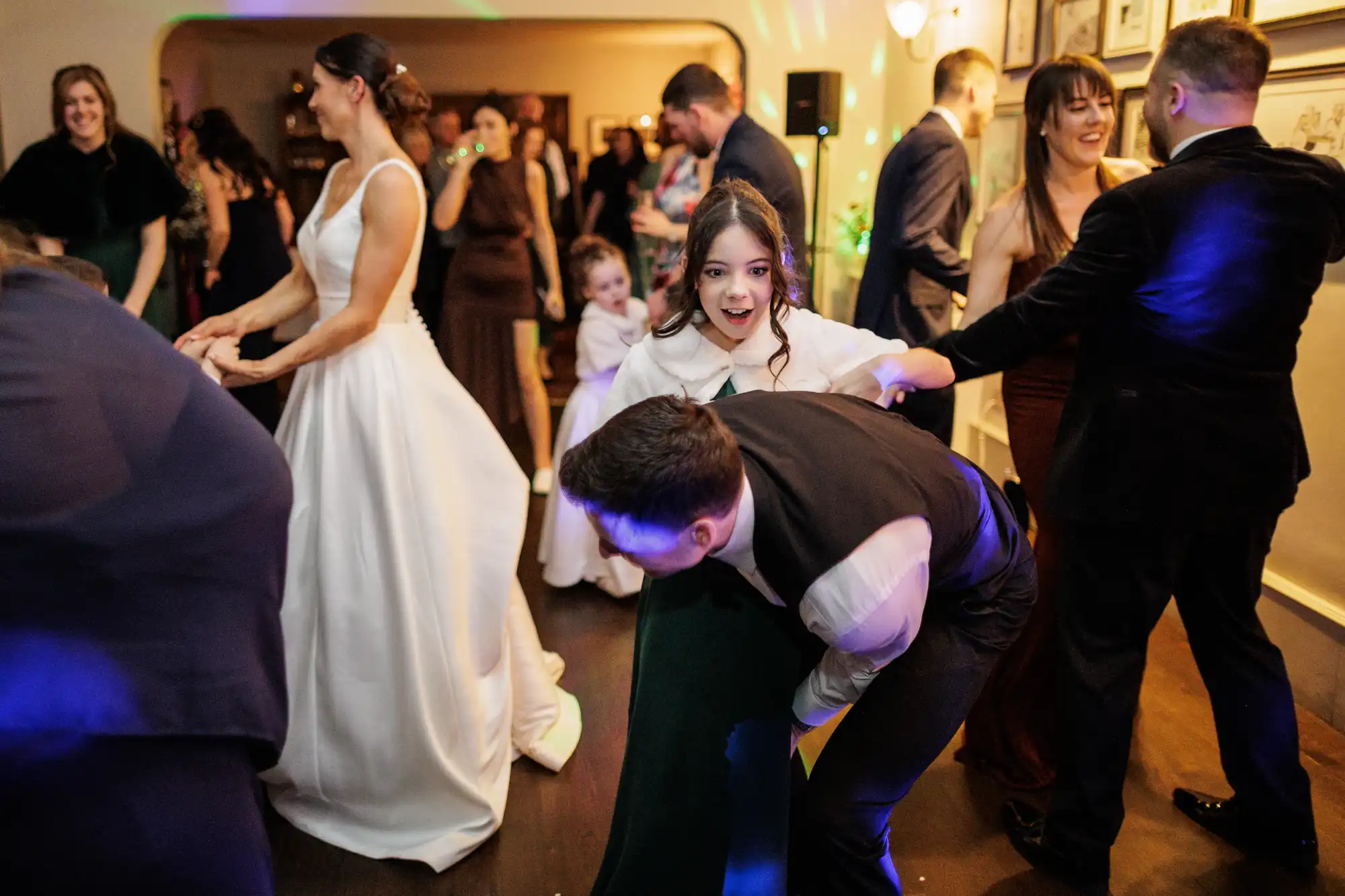 A diverse group of people are dancing and celebrating at a party. A bride in a white dress is on the left, a young girl in a white dress with a green collar is in the center, and a man in a dark suit is on the right.