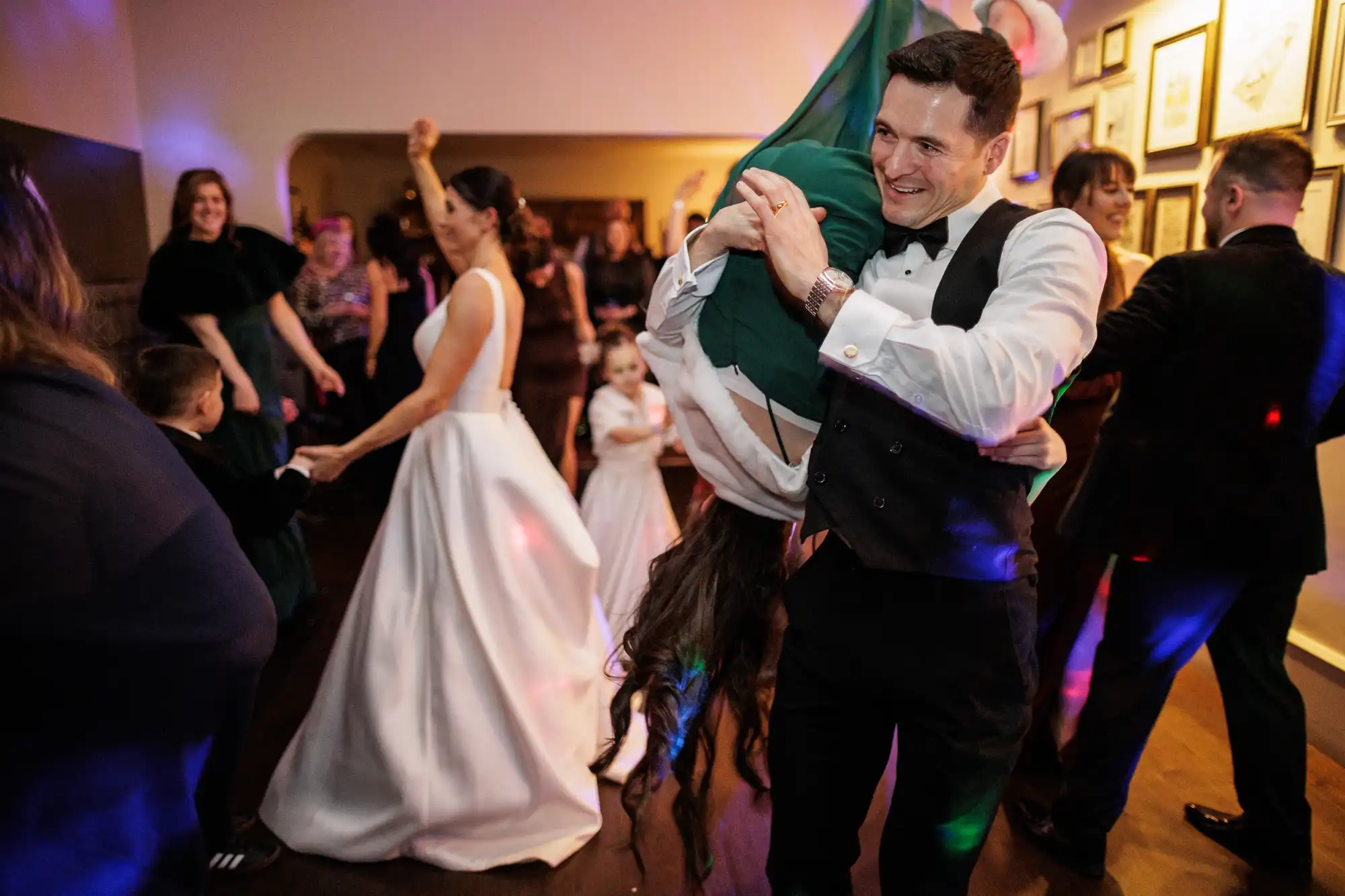 A groom in a tuxedo lifts a bride wearing a white gown upside down on a dance floor at their wedding reception.