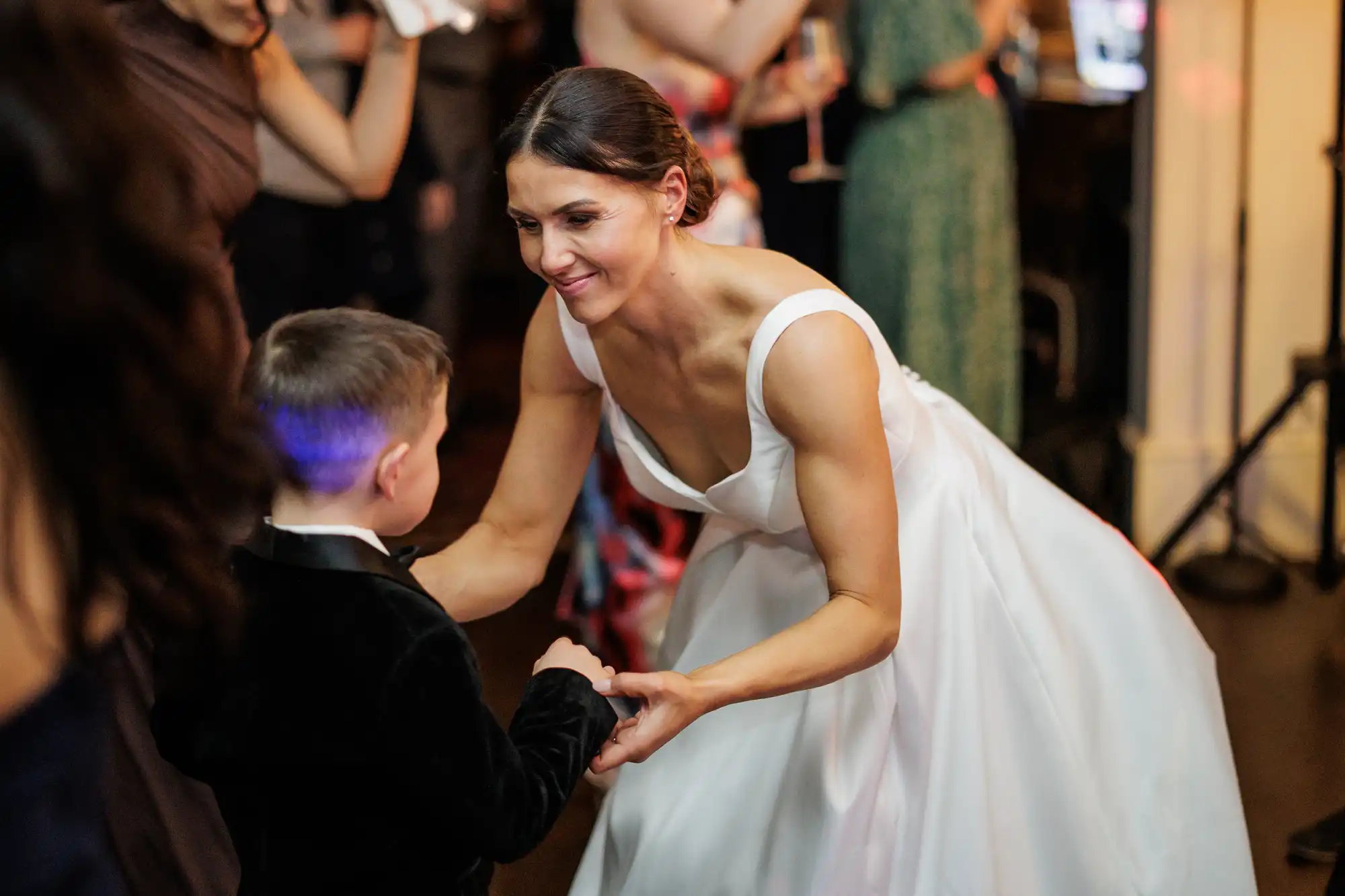 A smiling bride in a white off-shoulder gown bends down to hold hands and dance with a young boy in a black tuxedo at a wedding reception, with blurred guests visible in the background.