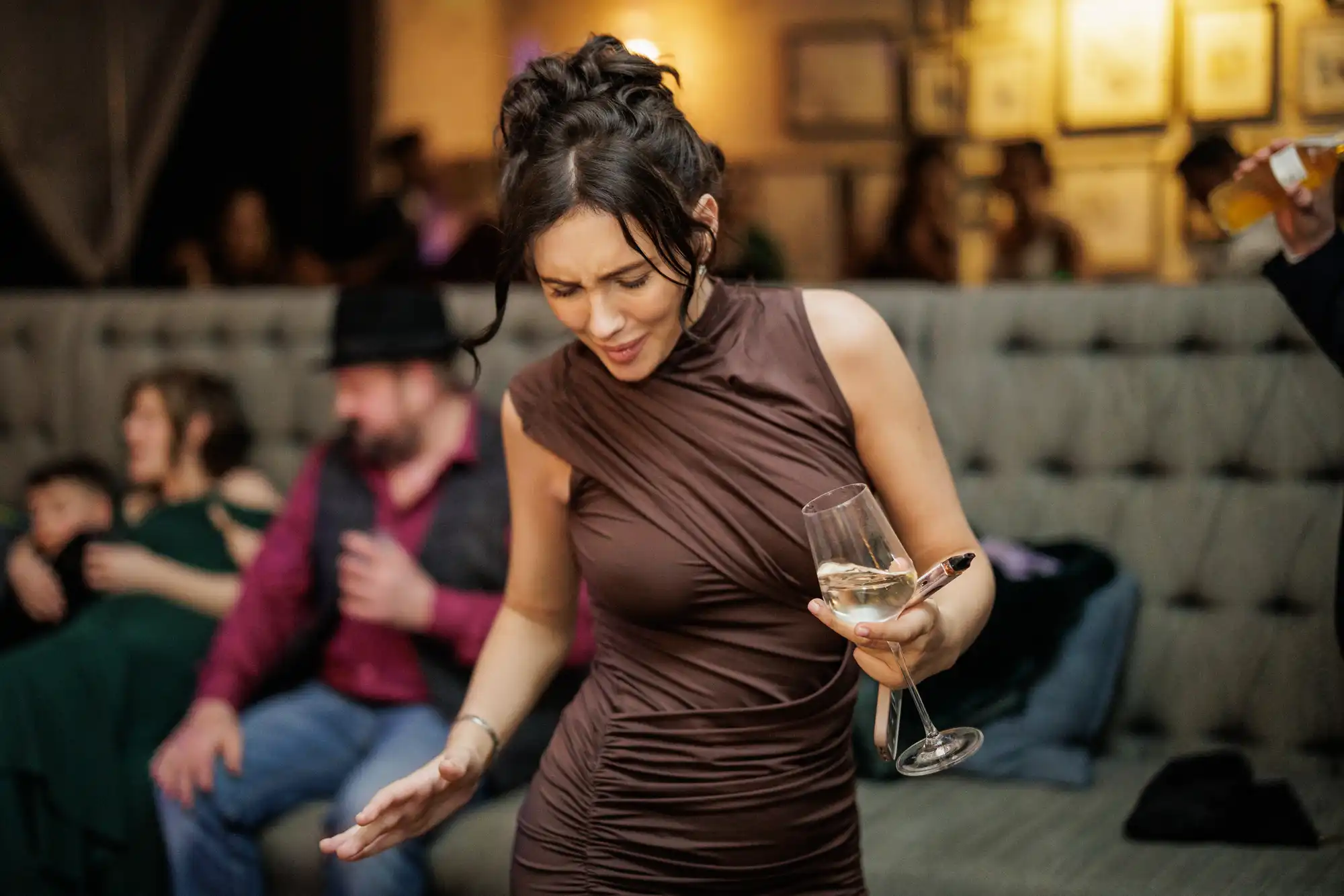 A woman in a brown ruched dress holds a wine glass, her head tilted down as if mid-conversation or dancing. People are blurred in the background.