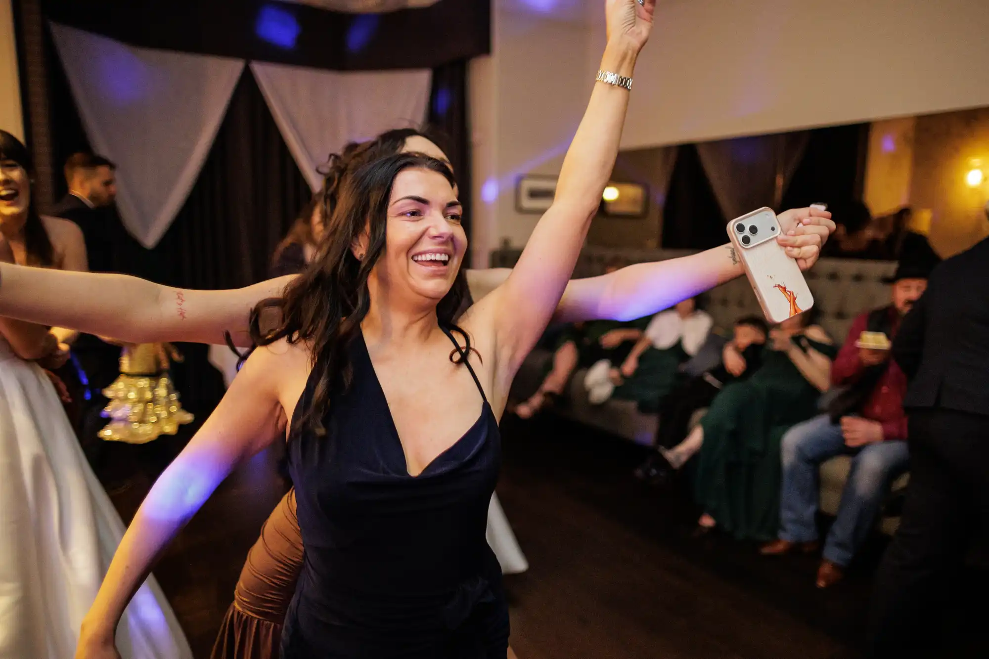 A woman in a dark blue dress laughs with her arm raised holding a phone. Other partygoers are visible in the background.