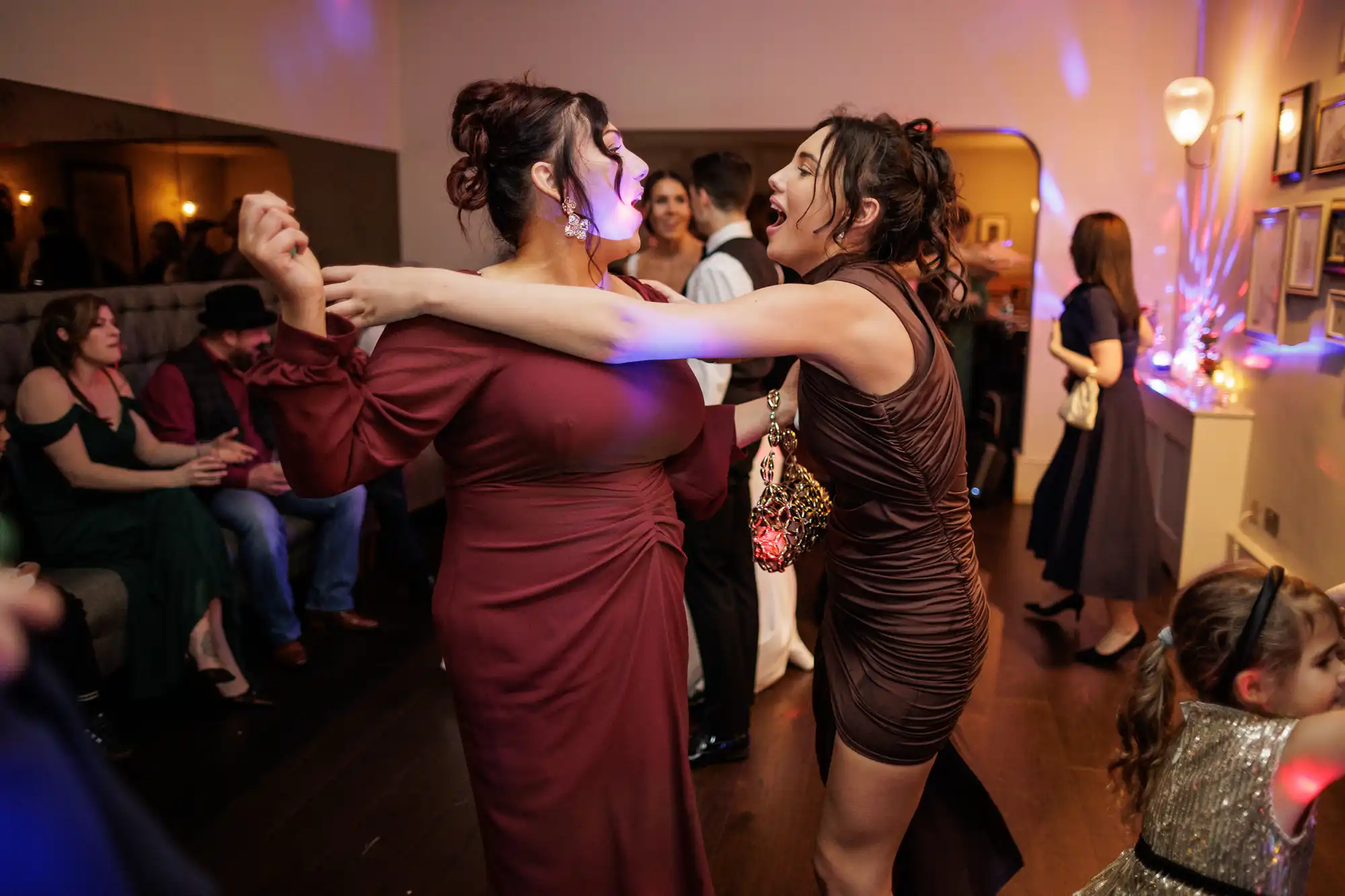 Two women in formal dresses dance closely, laughing and embracing. Lights reflect off their outfits. Other guests are visible in the background at a party.