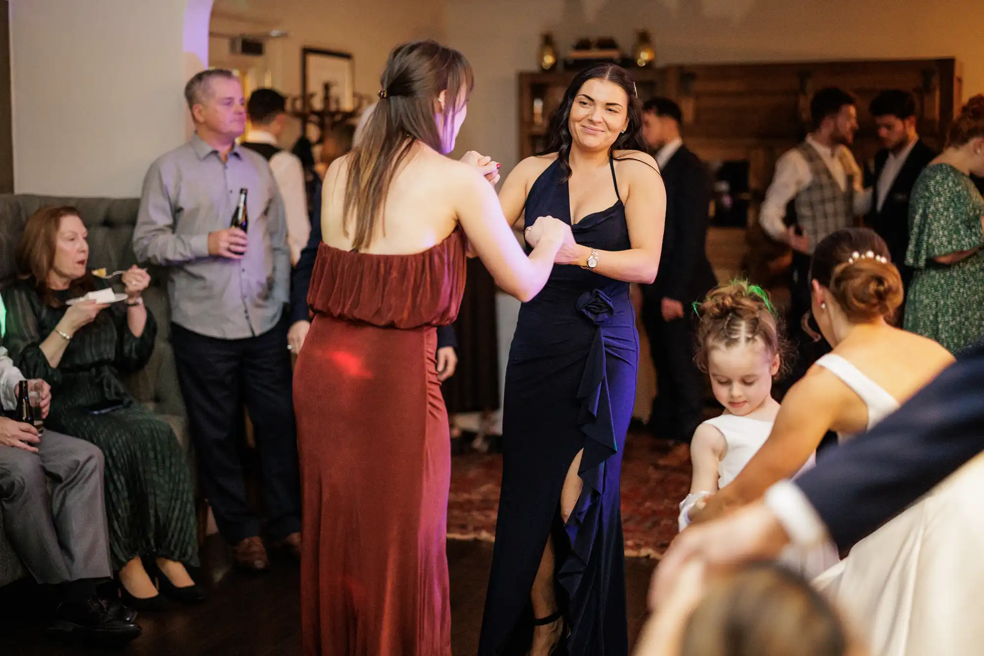 Two women dance at a reception. A woman in a rust-colored dress dances with a woman in a navy blue dress. A little girl in a white dress watches them.