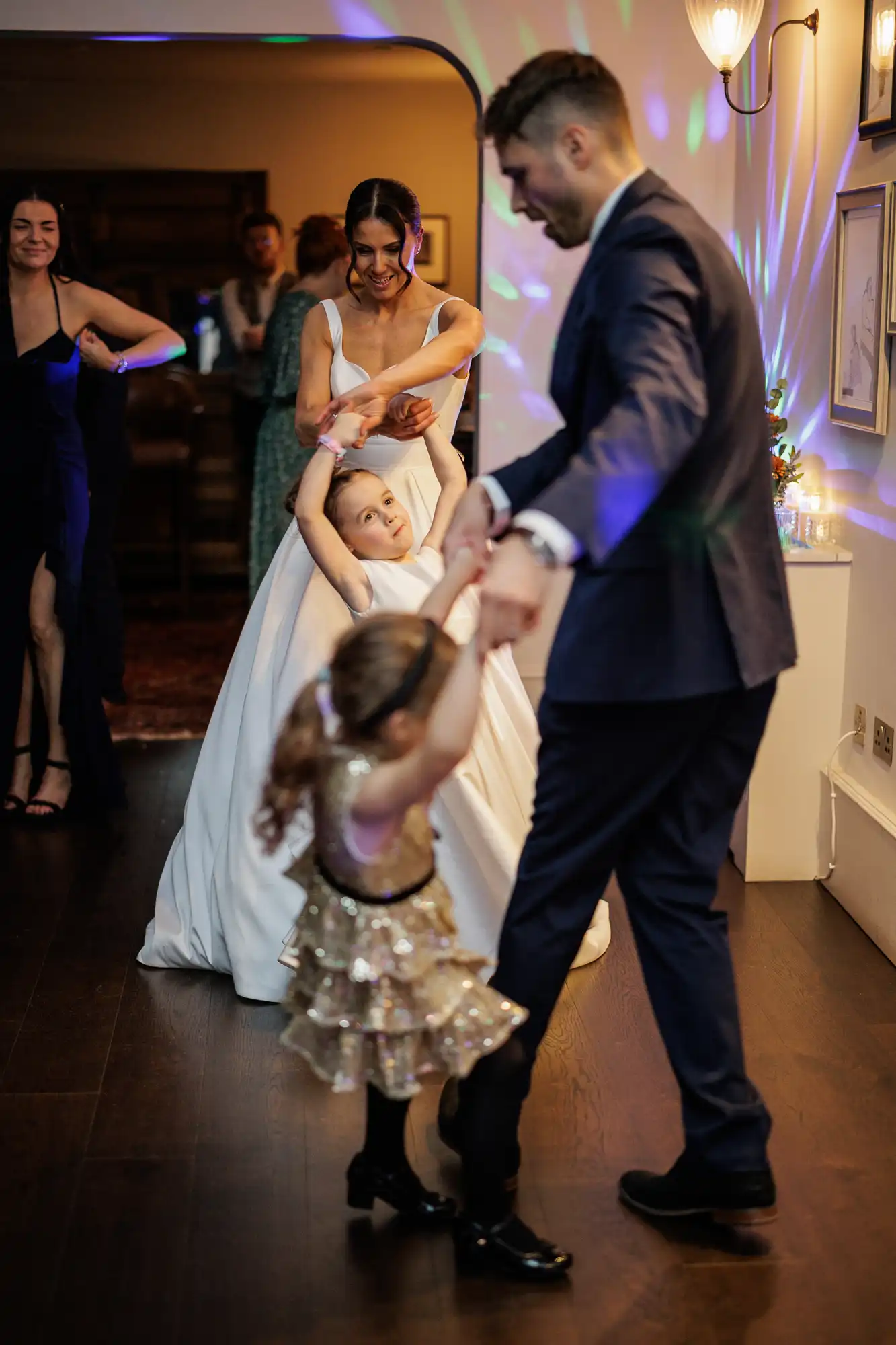 Bride and groom dance with two flower girls, one in a sparkly dress, at a wedding reception. Colorful lights reflect on the dance floor.