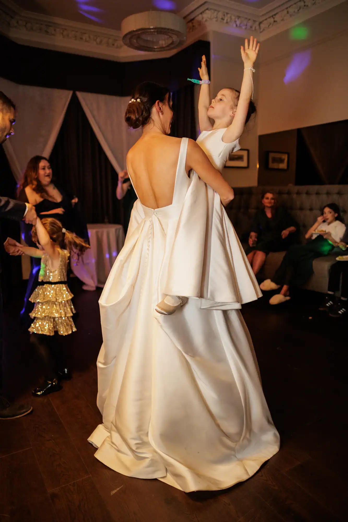 Bride holds a young girl in a white dress with arms raised, dancing at a reception. Another girl in a sparkly dress dances in the background.