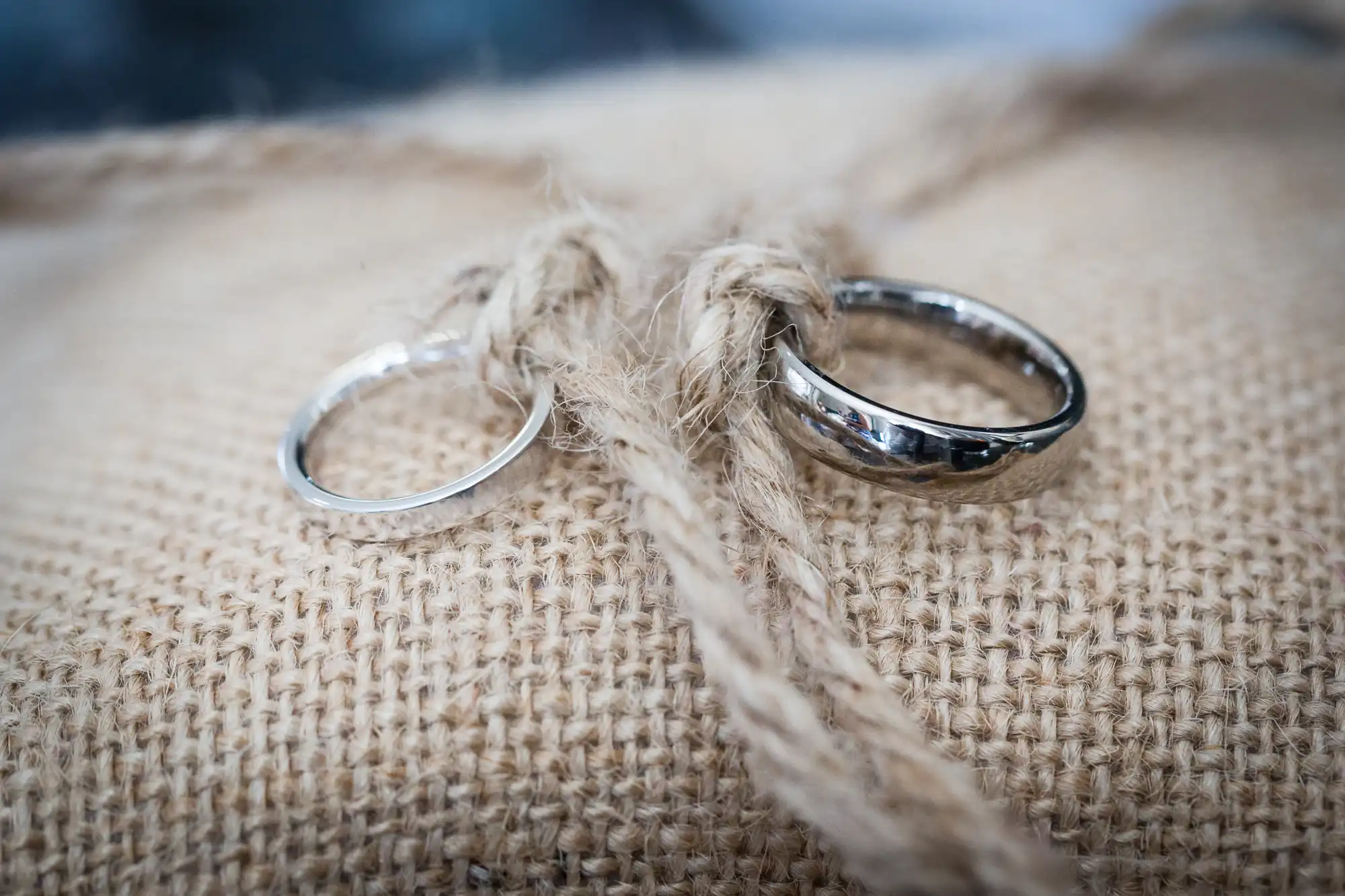 Two silver wedding rings are tied together with rustic string on a piece of coarse, light brown hessian fabric. The rings rest side by side, with one being slightly thicker and more reflective than the other, symbolising a wedding or commitment.