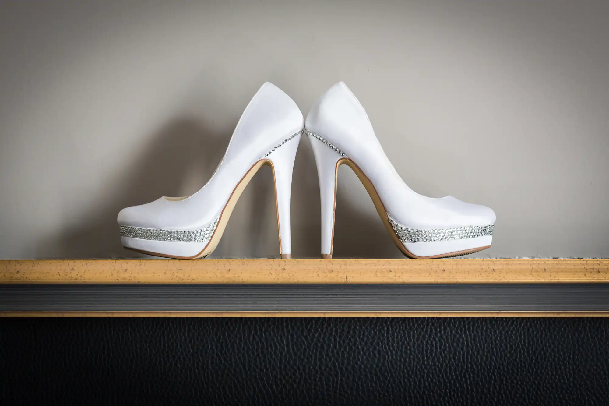 Two elegant white high-heeled shoes with silver rhinestone bands near the soles are placed sole to sole atop a wooden ledge against a plain grey wall, with the heel tips touching the surface.