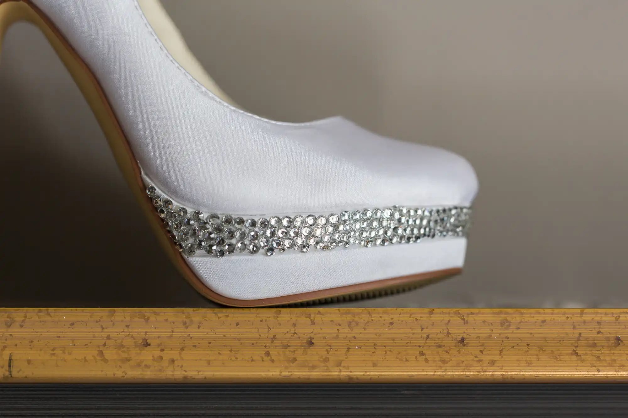 A close-up of a white high-heeled shoe with a platform toe. The platform is decorated with a band of small, sparkling diamanté. The shoe is resting on a textured, light brown surface.
