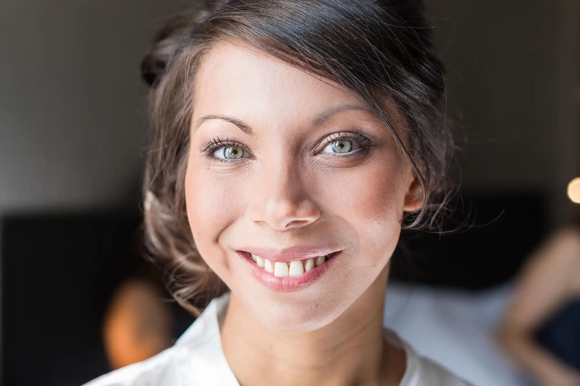 A woman with light eyes, brown hair styled up, and subtle make-up smiles warmly at the camera. She is wearing a white top, and the background is softly blurred, highlighting her bright, friendly expression.