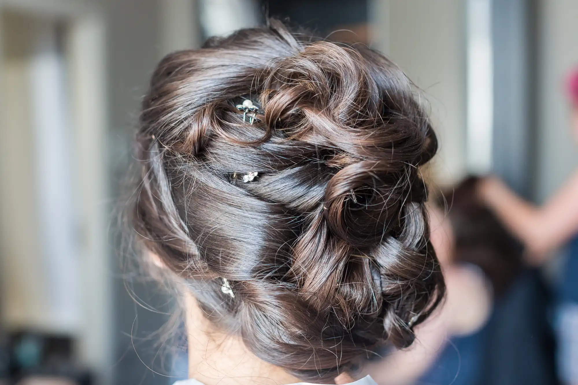 A close-up of the back of a woman’s head shows brown hair styled in an elegant updo with loose waves and twists. Small decorative hairpins with silver and crystal details are placed throughout the hairstyle. The background is softly blurred.