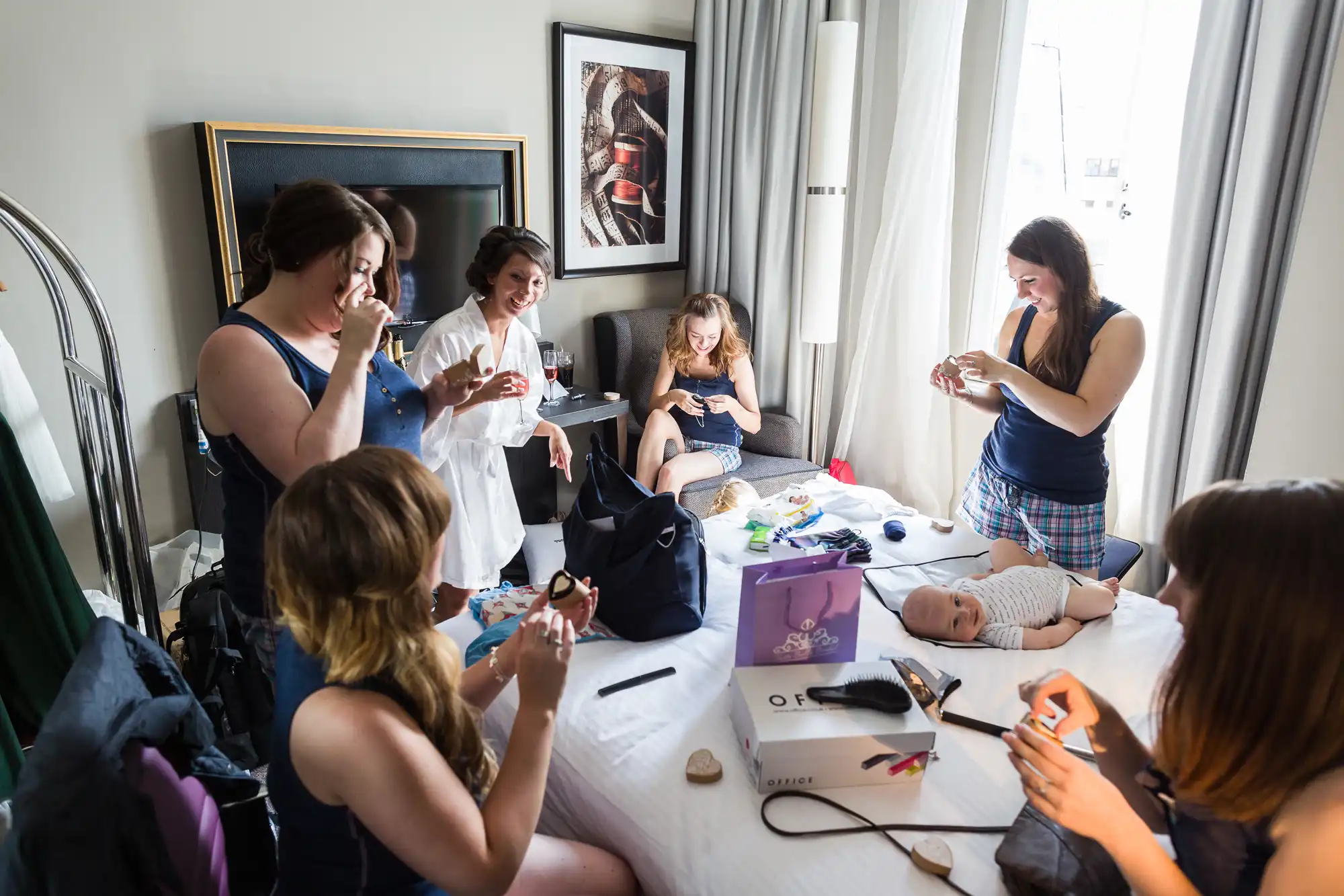 Six women in casual clothes sit and stand around a hotel room, smiling and applying make-up. A baby lies on the bed amid beauty products and bags. The room is bright with natural light from a window, and modern art hangs on the wall.