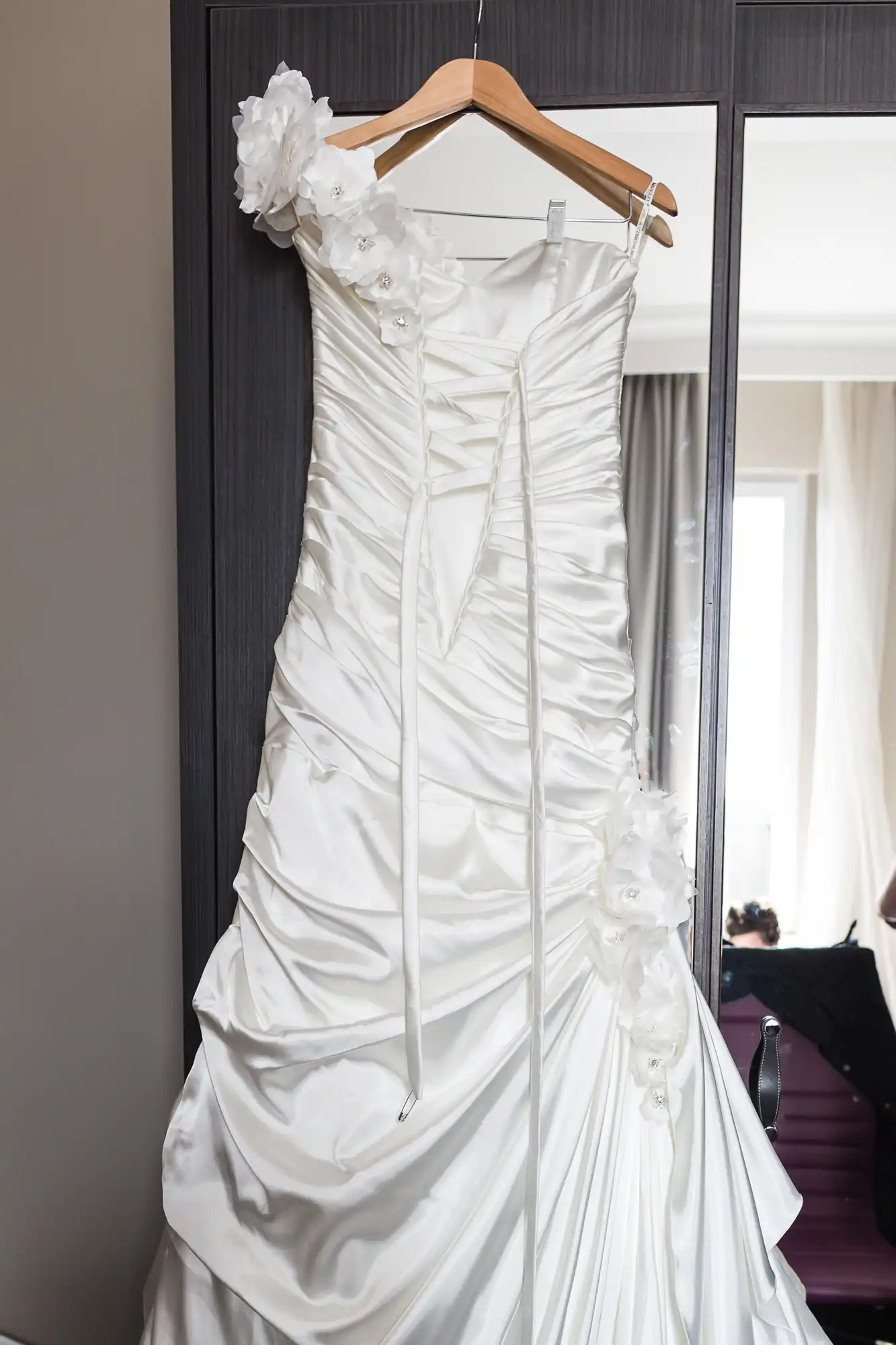 A white satin wedding dress hangs on a wooden hanger against a dark wardrobe. The dress is ruched with a fitted bodice, has floral embellishments on one shoulder and hip, and features ribbons at the front. A window and curtains are visible in the background.